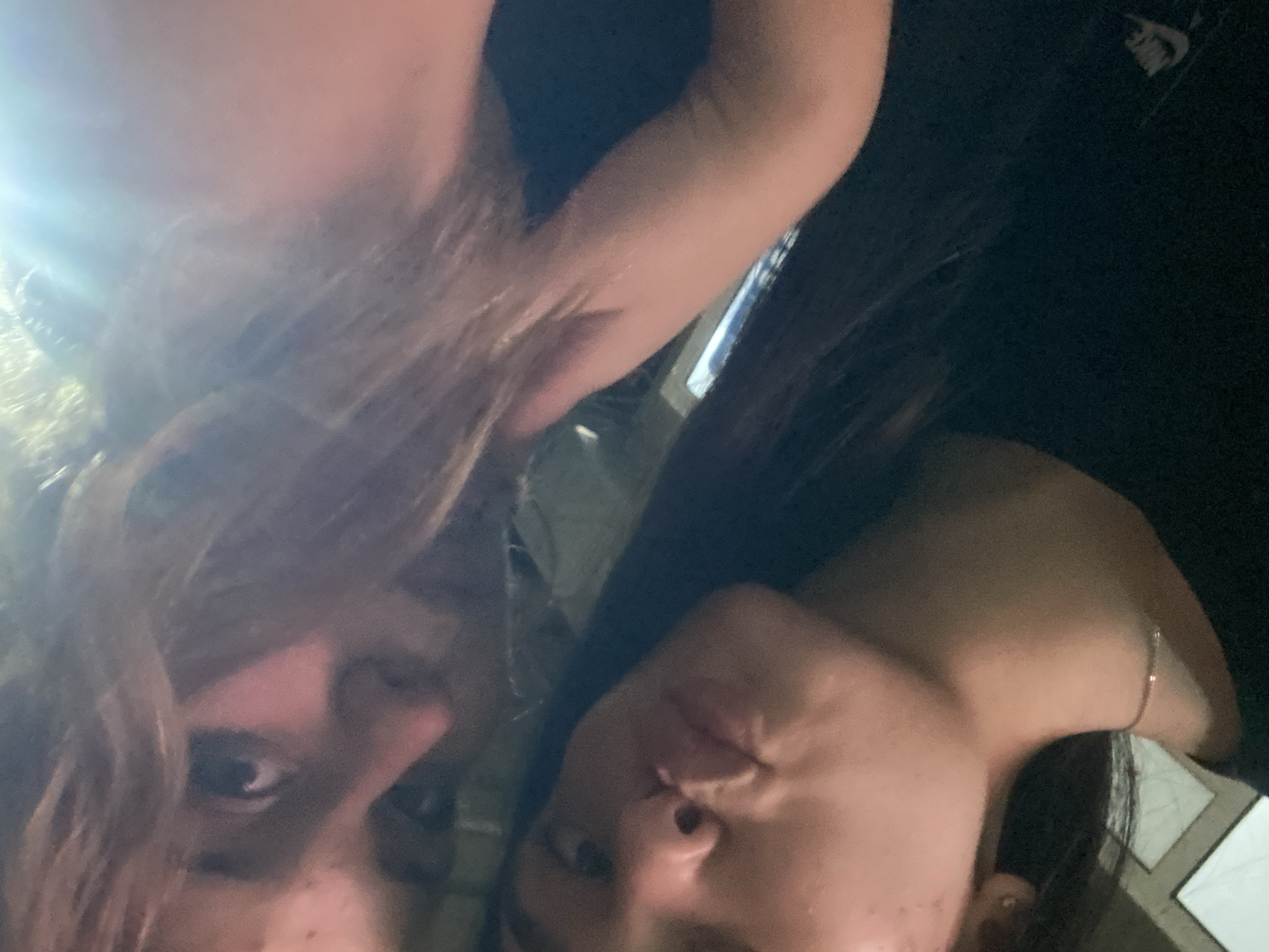 Two young women are shown in a close-up selfie. The woman in the foreground, on the left, is looking at the camera with her lips puckered. Her dark hair is visible, as is a thin silver necklace around her neck. The woman behind her, on the right, has lighter-colored hair that partially obscures her face. A bright light source illuminates the upper right portion of the image, creating a lens flare effect. Both subjects are wearing dark clothing. The background is dark and indistinct, with what appears to be a window or reflective surface visible in the lower left.