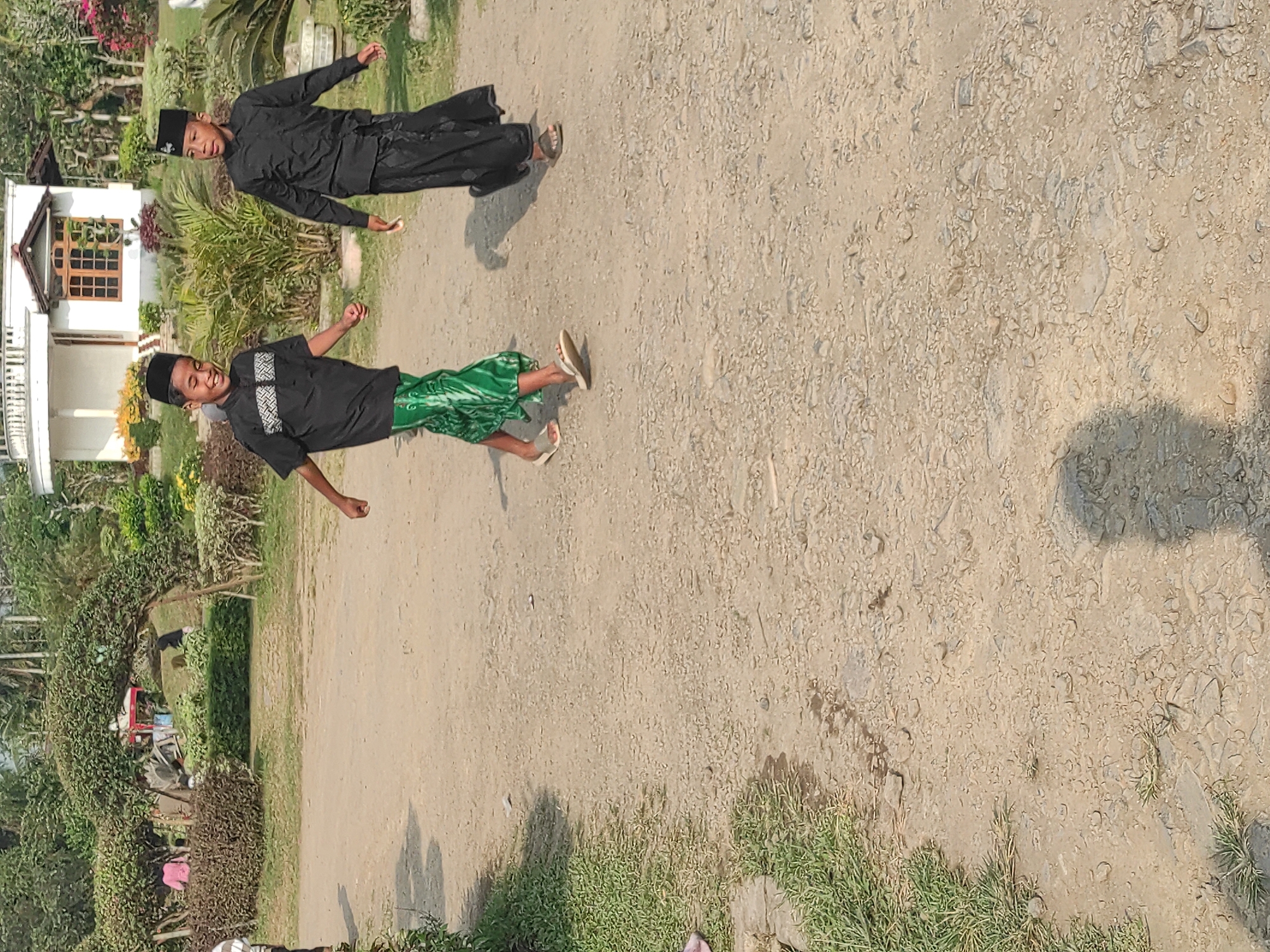 Two young boys wearing black long-sleeved shirts and black skullcaps walk on a dirt path in an outdoor park. The boy on the left wears a vibrant green sarong with a patterned design, while the boy on the right wears a black sarong. Behind them, a white building with a red-tiled roof is visible, surrounded by lush greenery, manicured bushes, and trees. Sunlight casts shadows on the path, indicating it is daytime. The scene suggests a casual outing in a well-maintained park.