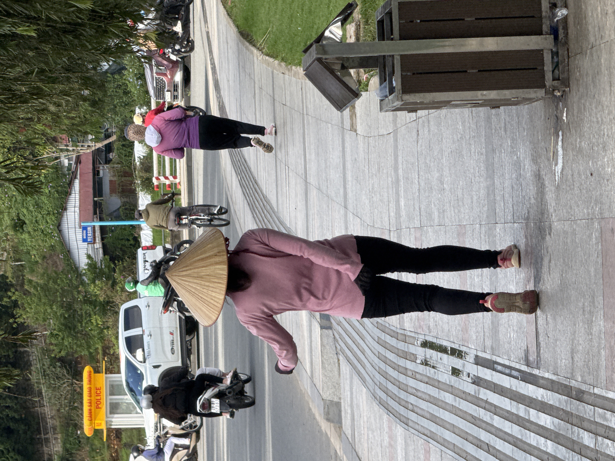 The image depicts an urban street scene in Da Lat, Vietnam, specifically featuring a section of Yersin Street, as indicated by the "ĐƯỜNG YERSIN" sign.
In the foreground, a person viewed from behind walks on a paved sidewalk. This individual wears a long-sleeved pink top, black pants, and a traditional conical straw hat (nón lá), with their left arm slightly extended and bent at the elbow, and their right hand near their hip. They wear two-toned sneakers, one pink, one beige. The sidewalk features a distinct pattern of parallel raised grey strips on the left, and smoother grey pavement on the right, with a small puddle visible on the patterned section. To the immediate right of the sidewalk, a manicured grassy area is present, bordered by a wooden trash receptacle with a slanted metal lid, containing visible refuse.
On the road to the left of the sidewalk, traffic is visible. A person on a bicycle, wearing a green/olive jacket, rides away from the camera. Behind them, a white pickup truck, identified by a "Ford" logo, is either parked or moving slowly. Next to the truck, a motorcyclist in a bright green jacket is also proceeding away from the camera. Further behind the truck, a yellow traffic police booth with "CẢNH SÁT GIAO THÔNG POLICE" signage is visible. To the left of the main subject, a motorcycle with two riders, a driver in a dark jacket and a passenger in light pants, is visible. The motorcycle's license plate reads "29-B1 573.63".
Further down the sidewalk, another person in a purple hooded jacket and black pants is jogging, moving in the same direction as the person in the conical hat. On the road next to them, a dark red SUV is visible, along with another motorcyclist in a red top and yellow helmet. The background features lush green hills with dense foliage and scattered buildings with red corrugated roofs. Overhead, tree branches with green leaves partially obscure the sky. The scene is well-lit by natural daylight.
