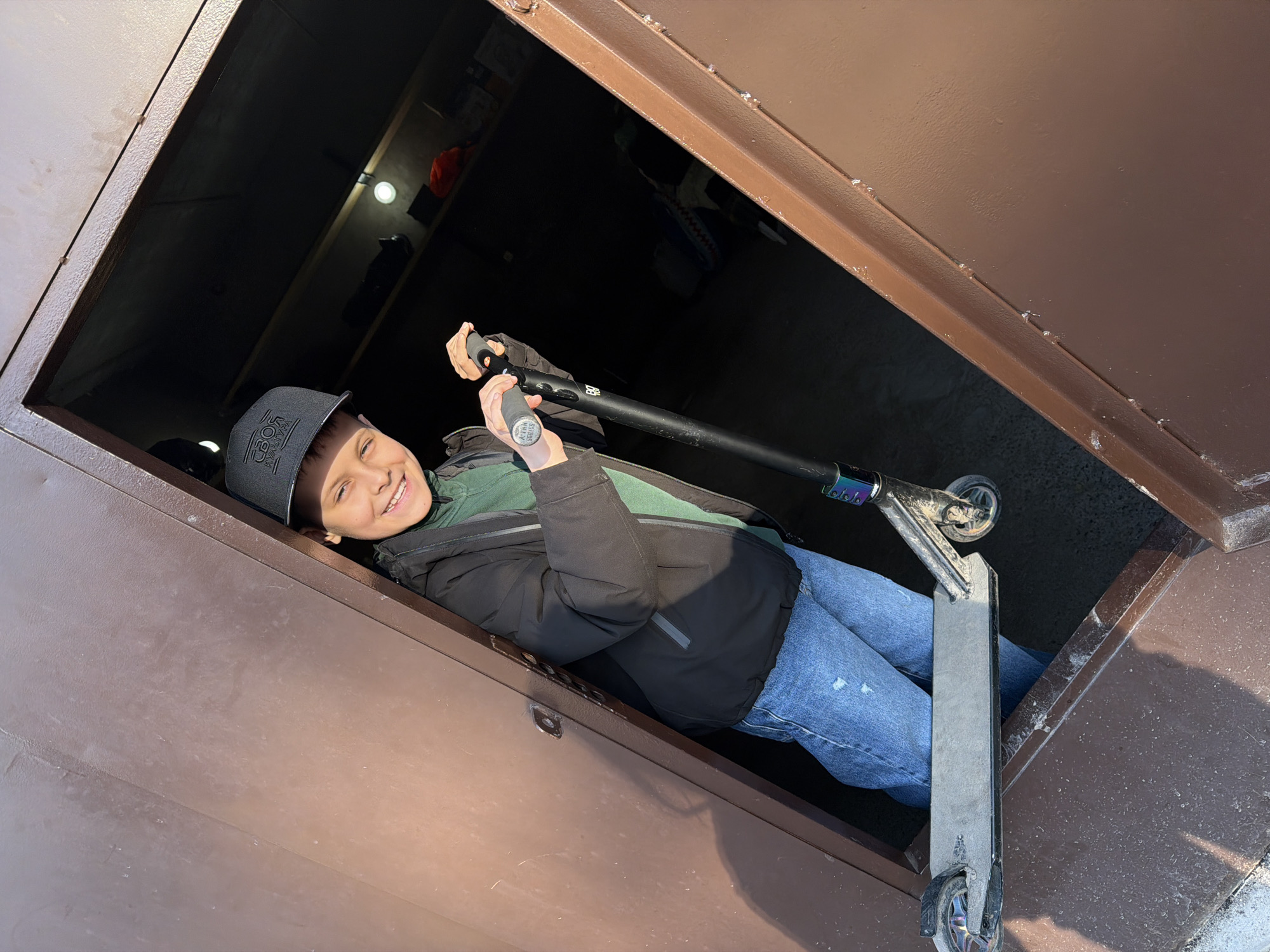 A young boy or teenager is depicted smiling broadly while emerging from a dark, enclosed metal structure, possibly a cellar or storage hatch, in what appears to be an outdoor setting in Votkinsk, Russia. The image is taken from a slightly upward angle, showing the boy positioned diagonally within the frame.
He is wearing a black baseball cap, a black puffer jacket over a green hoodie, and blue jeans. He is holding a black pro scooter with multi-colored accents on the handlebars and wheels. The scooter's deck is a dirty grey/silver.
Visible text on his baseball cap clearly reads "СВОЙ" (SVOY), meaning "OWN" or "ONE'S OWN" in Russian, with additional, less legible text below it. On the right scooter grip, the words "RIDER SERIES" are discernible.
The interior of the dark structure reveals a circular light source on the ceiling and faint shapes of stored items, including an orange bag and a blue, shark-shaped object. The exterior of the metal structure is a dull brown/bronze, showing some wear and possibly moisture marks. The bright sunlight creating distinct shadows suggests a clear, sunny day. The boy's expression conveys happiness or amusement.