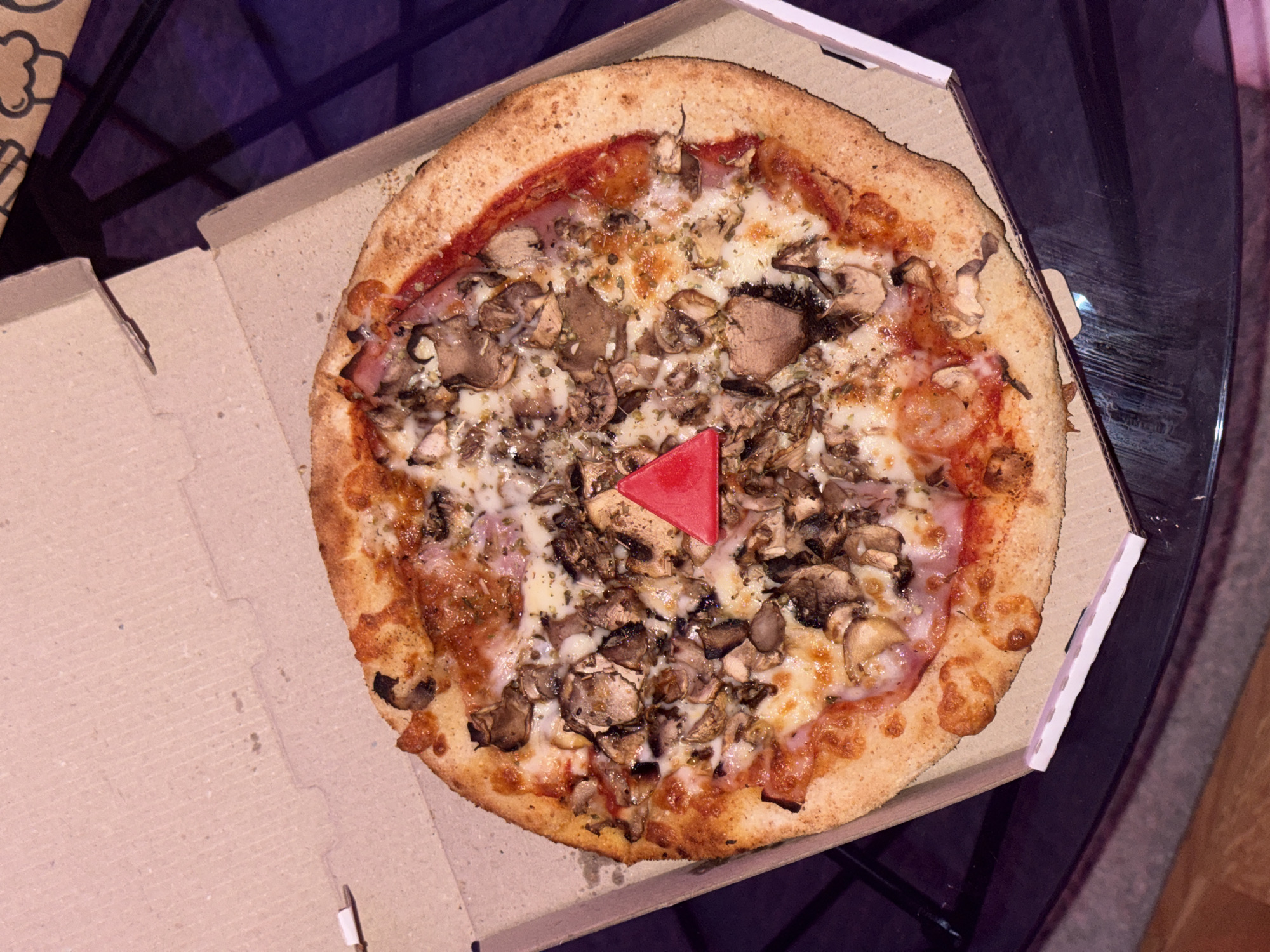 The image shows a pizza in a cardboard box, sitting on a dark glass table. The pizza has a thin crust, tomato sauce, cheese, ham, and is topped with a generous amount of mushrooms. A red, triangular plastic piece is placed in the center of the pizza. The pizza is inside a brown cardboard pizza box, with the lid partially open. The background is dark, with a glimpse of purple and brown suggesting an indoor setting. There is a design on another cardboard box, visible on the left. The location is unidentified.