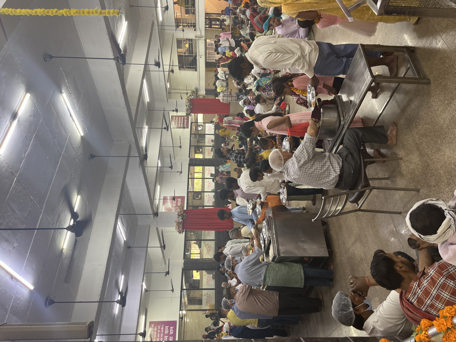 The image shows a large, crowded dining hall, likely a langar or communal kitchen, in a religious institution in Sector 12, Panchkula, India. It depicts a scene of food distribution and consumption, with a long line of people, mostly men, wearing simple clothing and head coverings, waiting to be served. Some are already seated at tables, eating their meals. There are several people serving food from large stainless steel containers. The hall has a high ceiling, white walls, and is brightly lit by fluorescent lights and ceiling fans. The atmosphere is one of organized chaos, with people moving and interacting, but with a general sense of order and purpose. There are signs on the walls in Hindi.  The walls feature large windows, and the scene is illuminated by natural light.  The floor is tiled and clean, and there are no visible signs of debris or dirt. Overall, the image captures a moment of communal gathering and food distribution in a religious context.