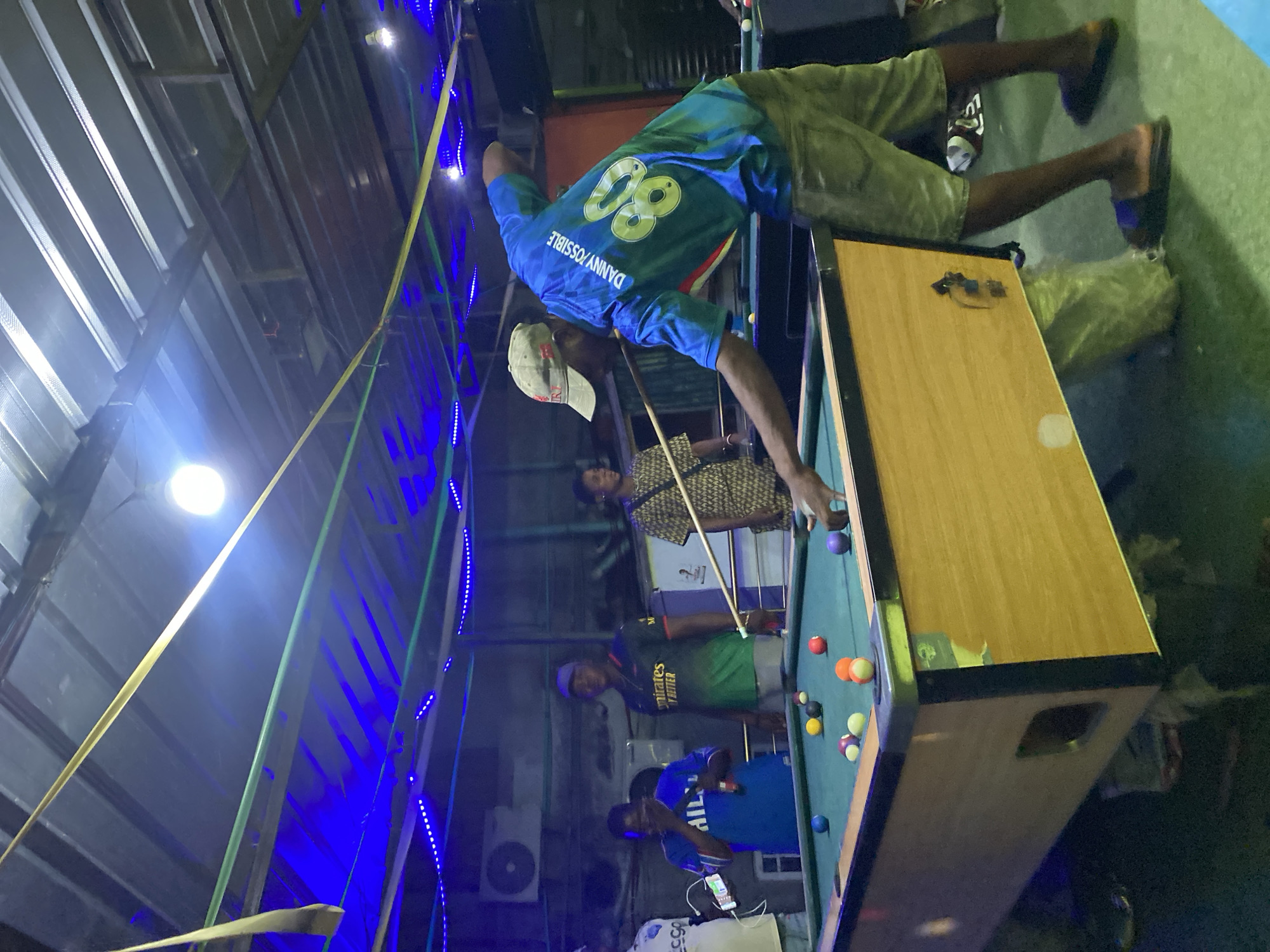 Captured in Ado, Nigeria, the image depicts several individuals engaging in a billiards game. An African man, wearing a blue t-shirt with the name "DANNY/POSSIBLE" and the number "80" emblazoned on the back, is poised to strike a billiard ball with a cue. Other men are gathered around the pool table, either observing the game or preparing for their turn. The room is illuminated by electric lights. A commercial air conditioner is installed in the background. The atmosphere is casual, indicative of leisure time. The pool table is surrounded by bystanders, creating a crowded ambiance.
