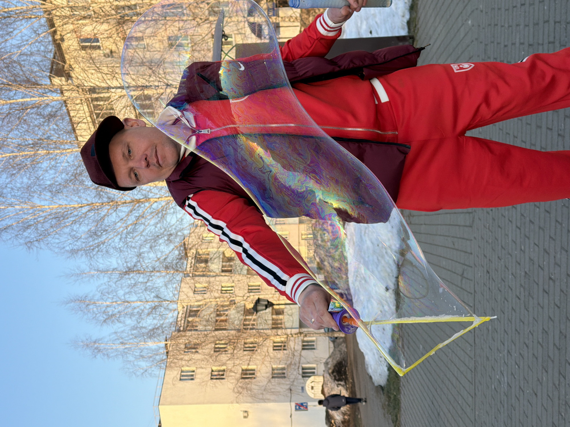 A man stands outdoors in Chaikovsky, Russia, holding a bubble wand and a large, iridescent soap bubble. He wears a red tracksuit with white stripes, a burgundy jacket, and a dark cap. The background shows a multi-story building with bare trees against a blue sky. Pavement is visible underfoot, and a small bird is on the ground behind him.