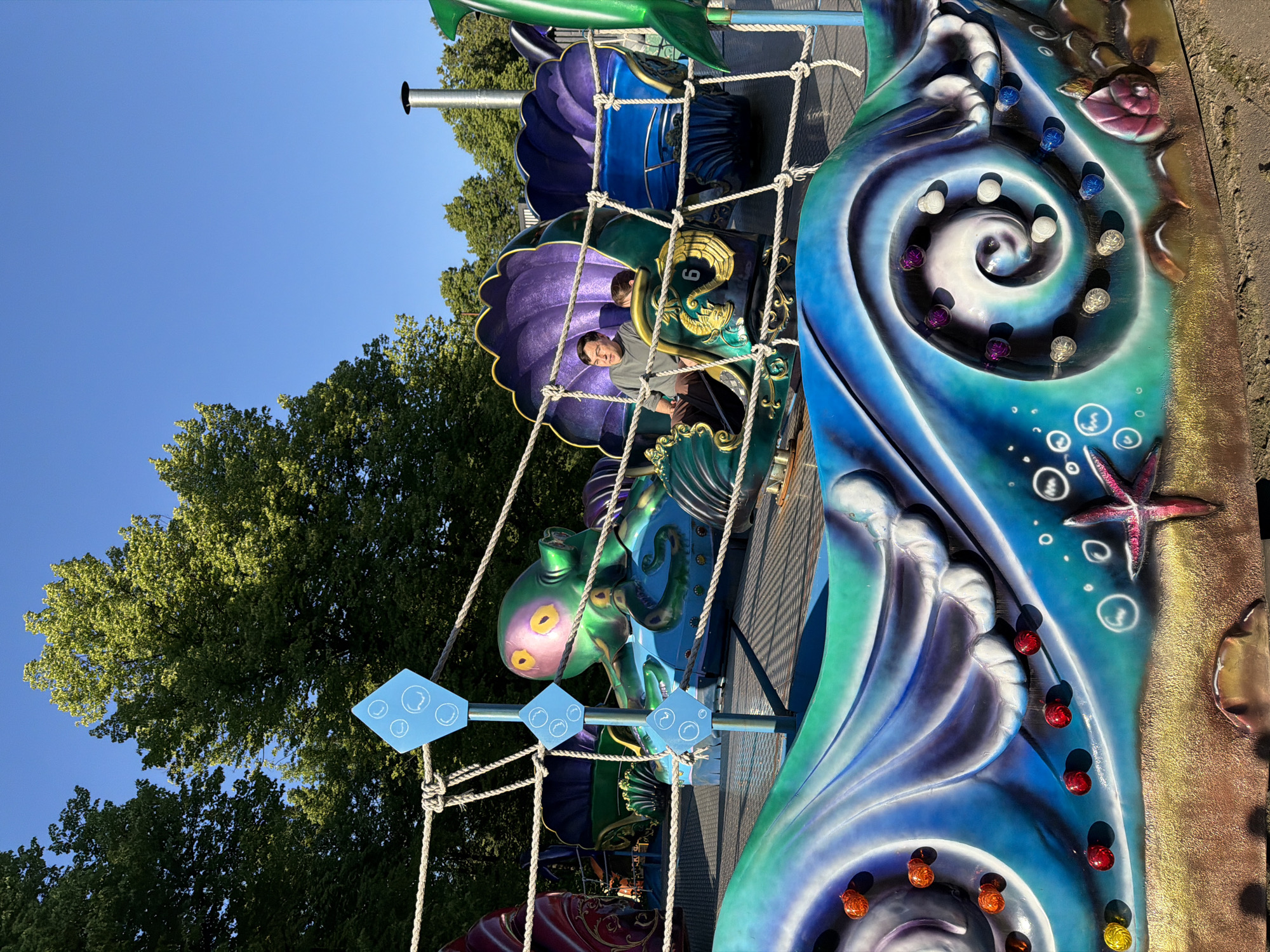 An outdoor amusement park ride with an aquatic theme is depicted under a clear blue sky, surrounded by dense green trees. In the mid-ground, an adult male with dark hair, wearing a grey long-sleeved shirt and dark pants, is seated in a purple, shell-shaped passenger cart, numbered "6". A child is partially visible next to him. The ride features multiple such carts and a large, green and purple octopus figure with yellow eyes. These elements are interconnected by light-colored rope netting.
The foreground displays the ride's decorative base, which is painted with stylized blue and green waves and a sandy seafloor design. Embedded in this base are various illuminated spherical lights in orange, red, yellow, purple, white, and blue. Painted details include white bubbles and a pink starfish on the "sand." A thin, blue vertical pole with three diamond-shaped blue signs, each showing white bubbles, stands in the foreground, supporting the rope netting. In the background, a tall, silver, cylindrical structure is visible on the right. This scene is consistent with an amusement park in Moscow, Russia.