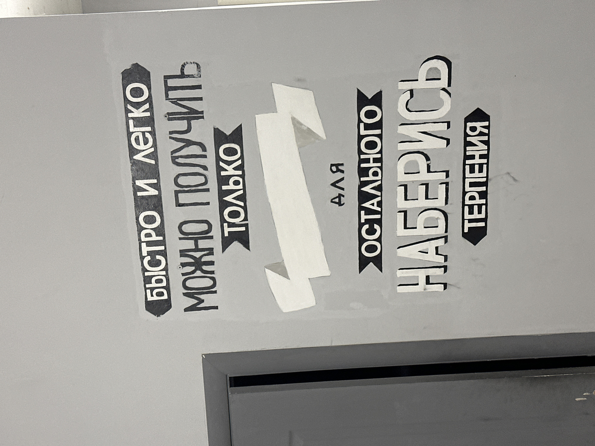 The image shows a sign in Russian that reads "Быстро и легко можно получить только для остального наберись терпения" which translates to "You can only get it quickly and easily, for the rest you need patience". The sign is attached to a gray surface. The lower edge of a dark gray object (possibly a door or panel) is visible below the sign. The location is implied to be Chaikovsky, Russia, based on the language of the sign.