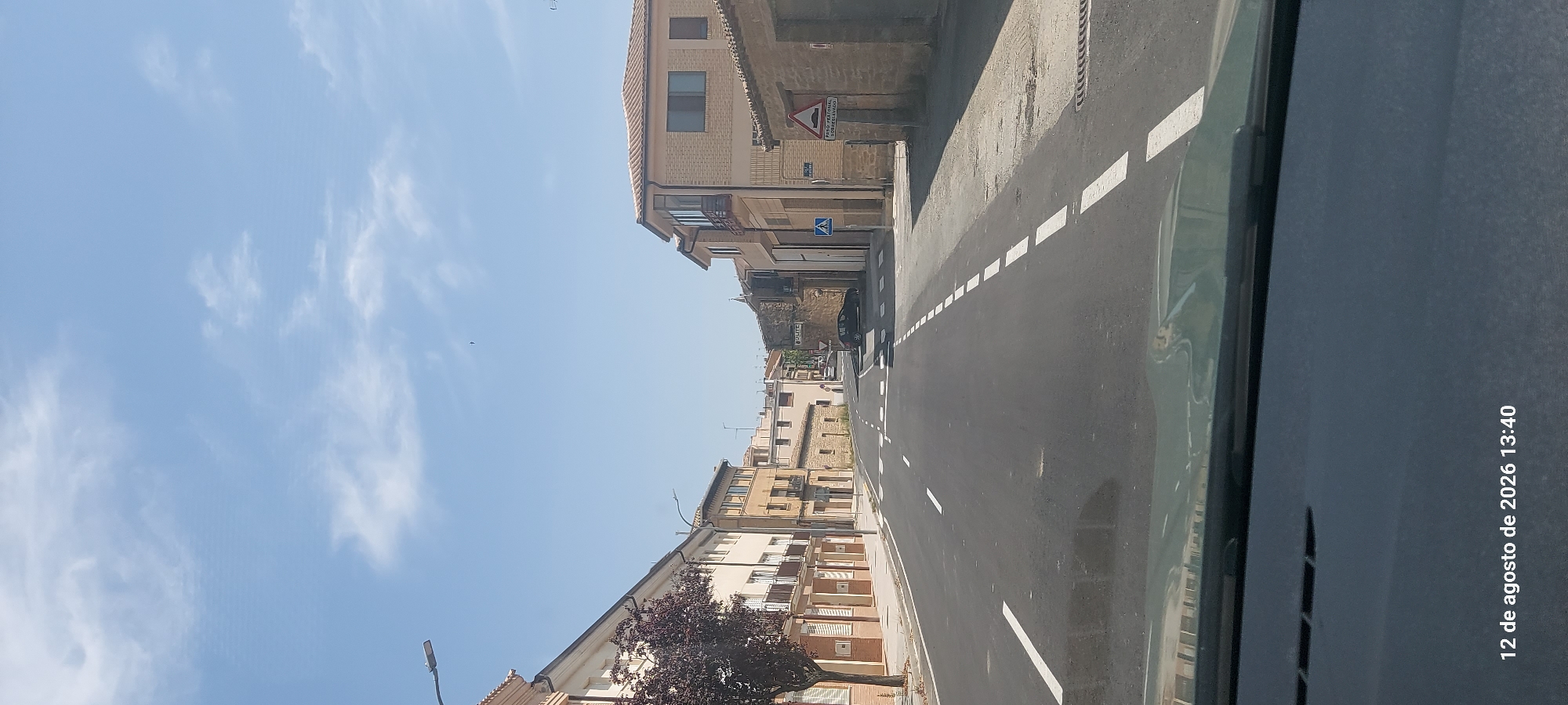 The image depicts a view of a street in Olite / Erriberri, Spain, likely taken from a vehicle. The street is paved with asphalt and has white lane markings, including a solid white line on the left and dashed white lines on the right, indicating the center of the road. A pedestrian crossing is visible mid-road. Buildings line both sides of the street. On the left, there are multi-story buildings with light-colored facades and balconies, and a tree with dark red foliage. On the right, buildings are constructed with stone and have tiled roofs. Traffic signs are visible, including a speed hump warning sign and a pedestrian crossing sign. The sky is blue with scattered white clouds. The date and time stamp at the bottom of the image indicates August 12, 2026, at 13:40.