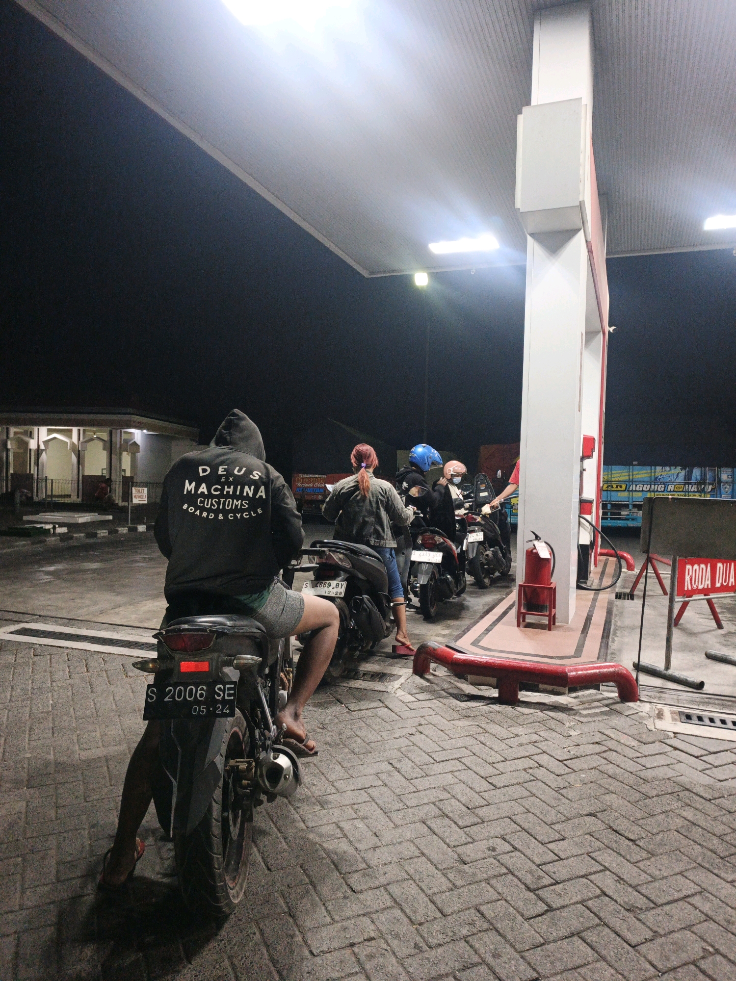 At a gas station in Ngrowo, Indonesia, at night, a person sits on a motorcycle facing away from the camera. They are wearing a dark hooded sweatshirt with "DEUS EX MACHINA CUSTOMS BOARD & CYCLE" printed in white on the back, and grey shorts. The motorcycle has a license plate that reads "S 2006 SE 05-24".
Several other motorcycles and people are visible in the background, near a gas pump. One person is wearing a blue helmet and another a pink helmet. A large blue and yellow truck is parked in the distance. The gas station has a white canopy supported by a large white pillar. A red fire extinguisher is mounted on a small red stand near the pump. A red sign with white text reading "RODA DUA" (which means "two wheels" in Indonesian) is visible to the right of the pump. The ground is paved with interlocking bricks. The overall lighting suggests it is nighttime, with artificial lights from the gas station and streetlights illuminating the scene. A building with arched windows and a mosque-like architectural style is visible to the left.