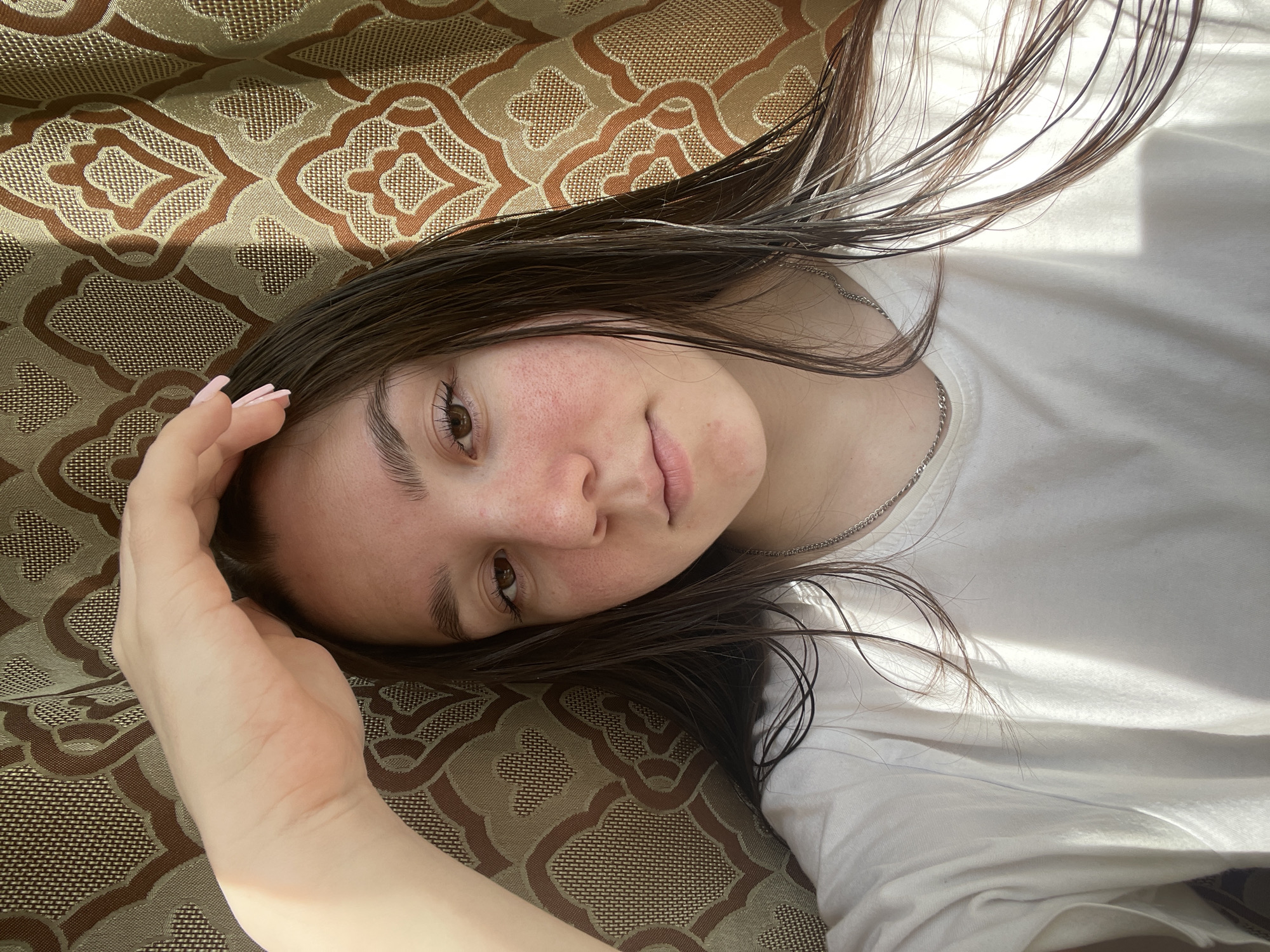 A young woman is lying down on a patterned brown and beige fabric surface, possibly a couch or bed. She is wearing a white t-shirt and a silver necklace. Her dark hair is spread around her head, and her right hand is raised near her forehead. Her skin appears slightly flushed. There is sunlight illuminating the scene. The image does not explicitly indicate the location of Ольховка, Russia.