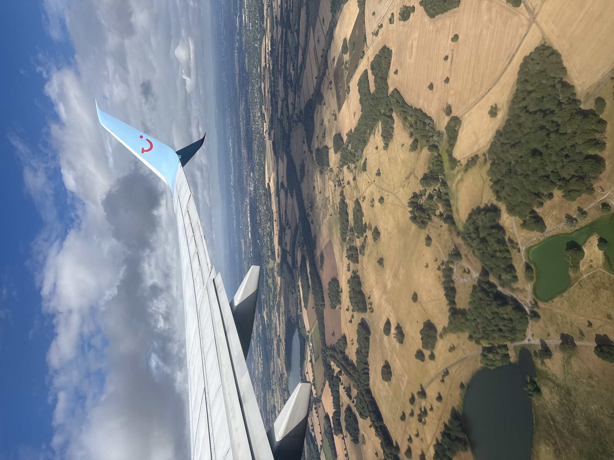This is an aerial photograph taken from an airplane window, looking down at a rural landscape. The dominant feature in the foreground is a section of an airplane wing, painted white and light blue, with the TUI airline logo visible on the wingtip. Below the wing, a patchwork of agricultural fields is visible, some appearing to be harvested with stubble, and others covered in dry grass or crops. Scattered clusters of trees, forming small woodlands, break up the fields. There are two bodies of water; a larger dark lake on the left and a smaller, bright green pond on the right. In the distance, a sprawling urban area can be seen, with buildings and infrastructure stretching towards the horizon. The sky is blue with scattered white and grey clouds. The photograph captures a view of the countryside, potentially near Knutsford, United Kingdom, given the presence of agricultural land, woodlands, and a distant urban center.