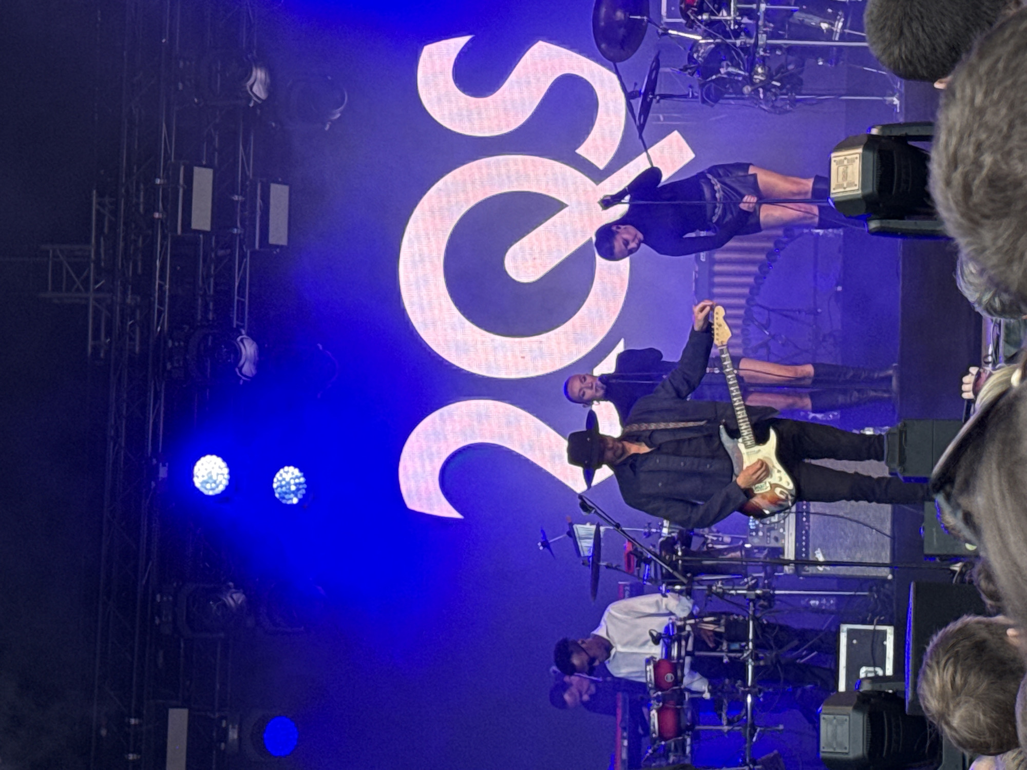 A band is performing on a stage under blue lights. The stage has a large, illuminated sign that says "20s" in a stylized font. A guitarist, wearing a black jacket, hat, and black pants, is in the foreground, holding a white guitar. A drummer, wearing a white shirt, is behind him, playing a drum kit. Several band members are visible on stage, and there are lights and audio equipment around them. The audience is visible in the foreground, with their heads and shoulders showing, suggesting the photo was taken from within the crowd. The lighting suggests it is nighttime, and the event is likely an outdoor concert given the presence of the audience in the foreground. The overall mood appears energetic and exciting, typical of a live music performance. The visible text is "20s".