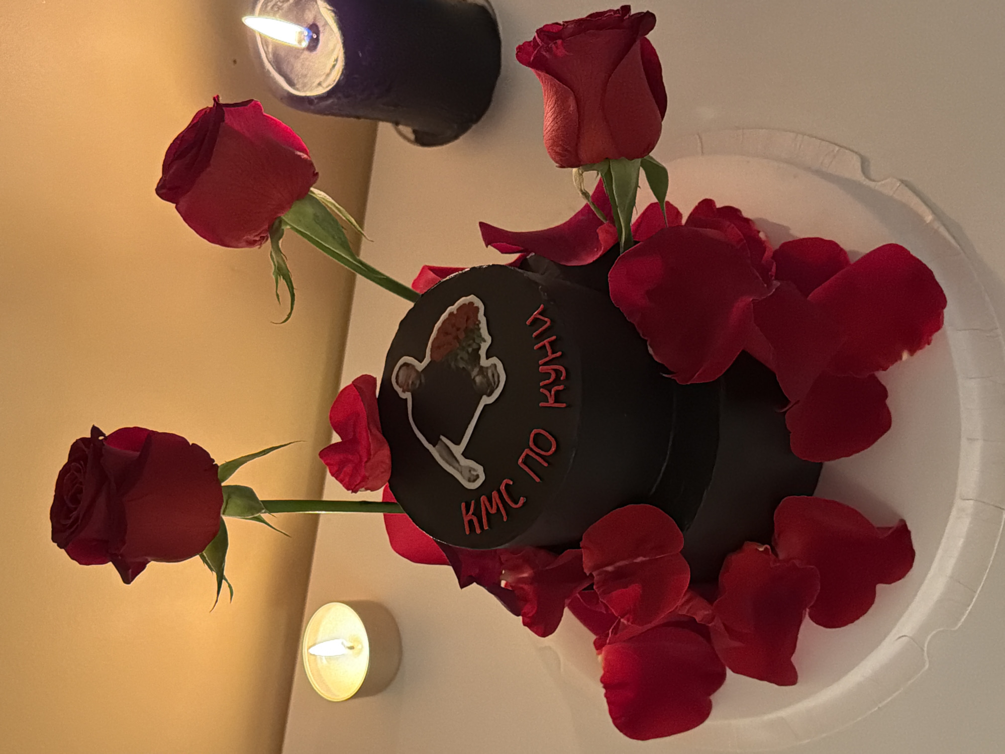 A still-life image shows a black, tiered cake decorated with red roses and scattered red rose petals. The cake has lettering in red on its side and a picture affixed to the top. The scene is illuminated by two candles, one near the top and another on the bottom left. The cake sits on a white, fluted-edge plate. The lighting suggests an indoor setting. The image doesn't provide enough information to ascertain a location in Tel-Aviv, Israel.