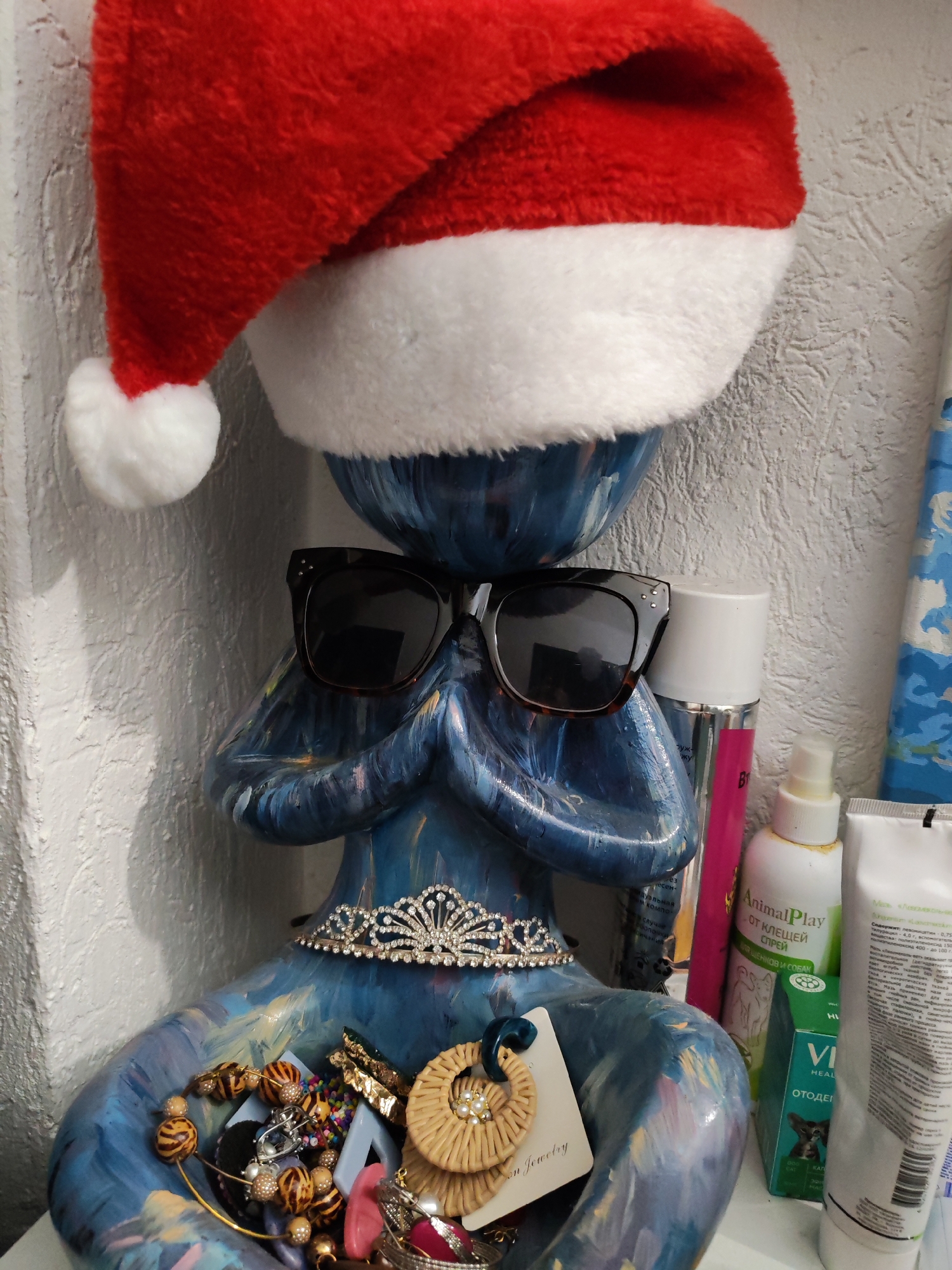 A blue figurine wearing a Santa hat and sunglasses sits among various items. The figurine also has a decorative tiara and holds jewelry in its bowl-like "hands." The surrounding items include bottles and tubes of what appear to be personal care products. The products seem to include a spray for ticks, marked as "AnimalPlay", and a medication for ears labelled "VI HEAL ОТОДЕ". A tag marked "On Jewelry" is visible among the jewelry. The background includes a textured, light-colored wall. The location is Izhevsk, Russia, based on Cyrillic text on the products.