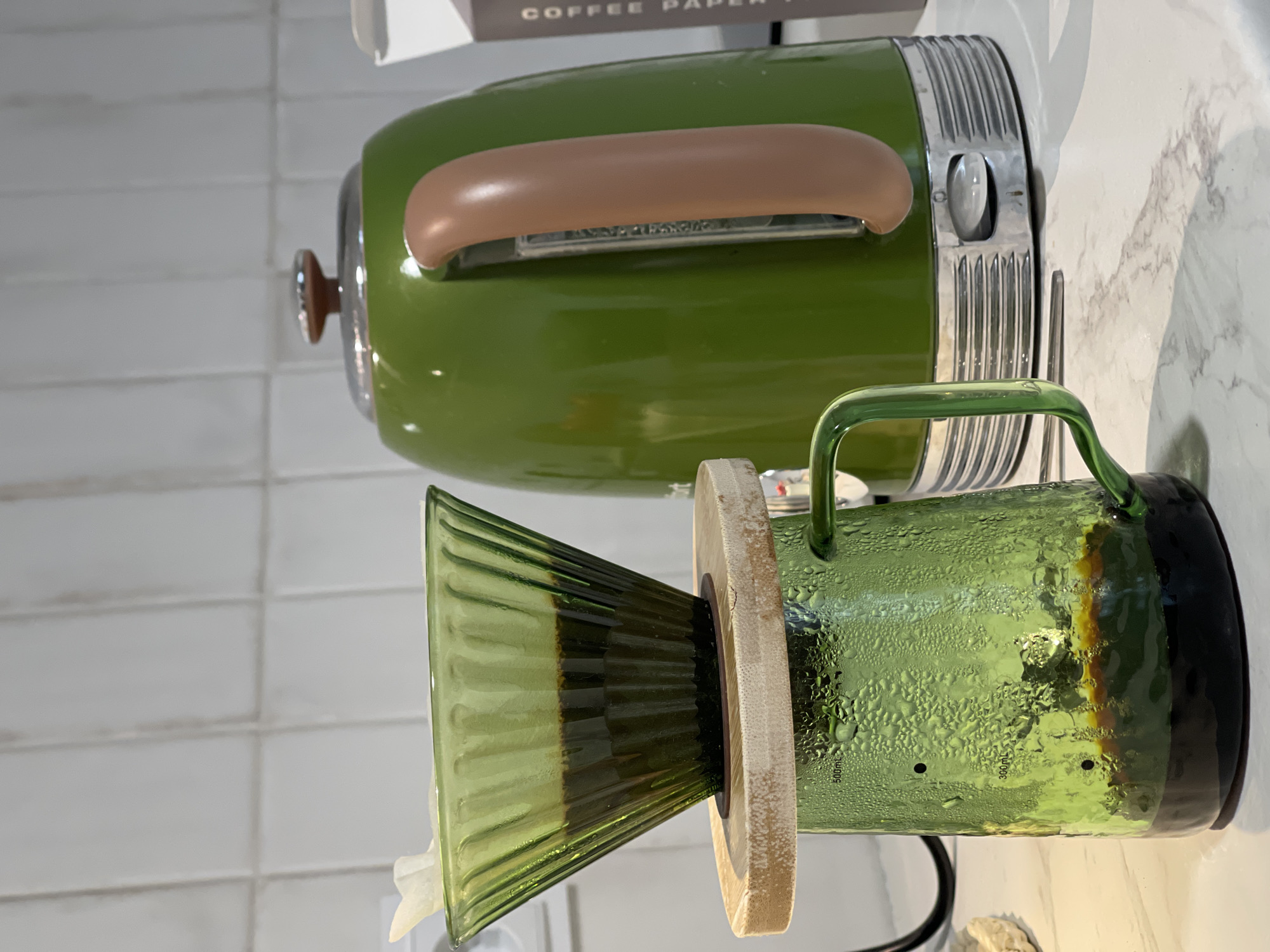 The image shows a green coffee setup. In the background, there is a green electric kettle with a brown handle, positioned on a white countertop. In the foreground, there is a green glass pour-over coffee dripper with a wooden ring and a glass carafe. A white coffee filter is visible inside the dripper. The setting appears to be indoors, possibly in a kitchen or coffee shop. There is no indication of the location being Izhevsk, Russia.