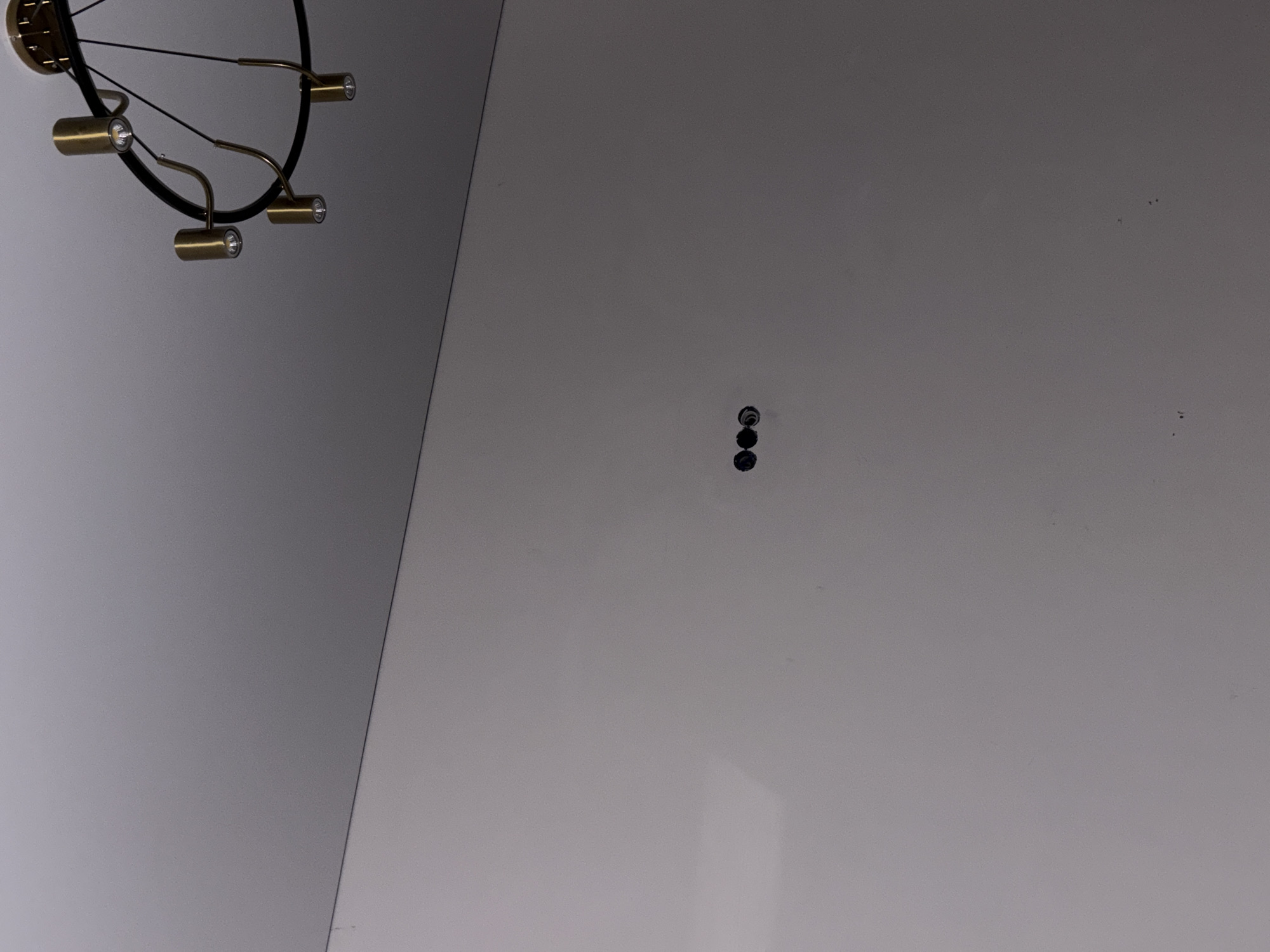The image shows a portion of a white ceiling with a circular black and gold light fixture in the upper left corner. In the center of the frame, closer to the right side of the image, are three small, dark holes arranged vertically in a line. The ceiling appears to be in Ембаево, Russia, based on implied location data; however, no information about people, actions, interactions, or other objects is evident in the image.
