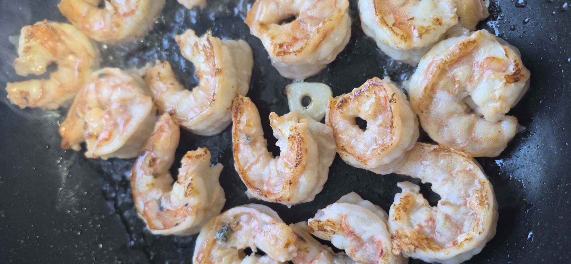 The image shows a close-up, top-down view of several large shrimp being cooked in a dark non-stick pan. The shrimp are pinkish-white with some golden-brown seared edges, indicating they are being sautéed or pan-fried. There is a small amount of liquid or oil visible in the pan, and wisps of steam rise from the shrimp, suggesting they are actively cooking. One slice of garlic is visible among the shrimp. The pan appears to be on a stovetop. There are no people or identifiable landmarks from Saint Petersburg, Russia, present in the image.