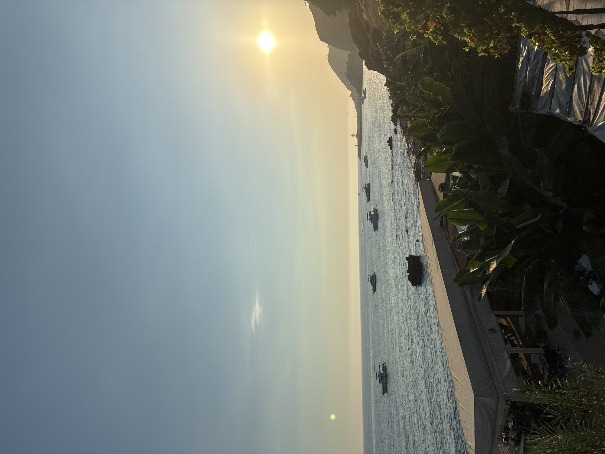 The image depicts a coastal scene in Alanya, Turkey, during what appears to be sunset. The sun is low in the sky, casting a warm, yellow glow and creating a bright haze on the horizon. The sea is calm with several boats of varying sizes anchored or moving slowly. The coastline is characterized by rocky shores and lush green vegetation, including large-leafed plants and some trees. In the foreground, a white, tent-like structure with visible support poles is situated along the waterfront, suggesting a possible outdoor dining or recreational area. The sky transitions from a light blue near the horizon to a deeper blue at the top of the frame, with a few faint clouds. The overall impression is one of a tranquil evening by the sea.