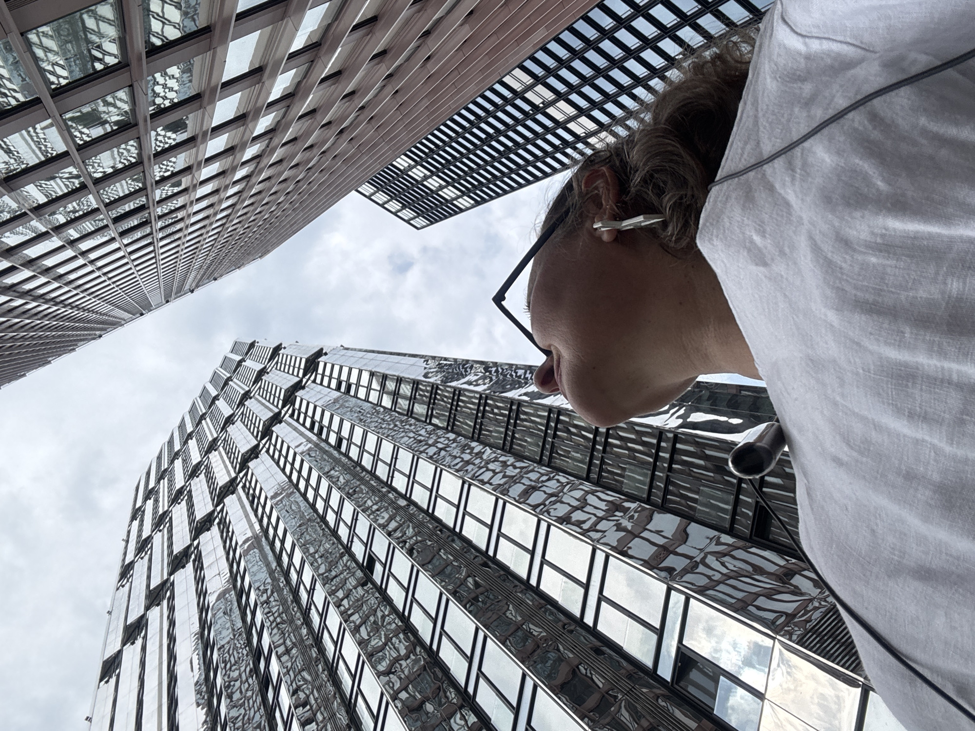 The image shows a low-angle shot looking up at two modern skyscrapers with reflective glass facades. The sky above is overcast. In the foreground, the back and shoulder of a person wearing a white t-shirt and headphones is visible. The person is wearing large, angular earrings and glasses. The skyscrapers are tall and imposing, with many windows reflecting the sky and surrounding structures. The overall impression is one of urban density and modern architecture. The scene is a street in the city of Khimki, Russia.