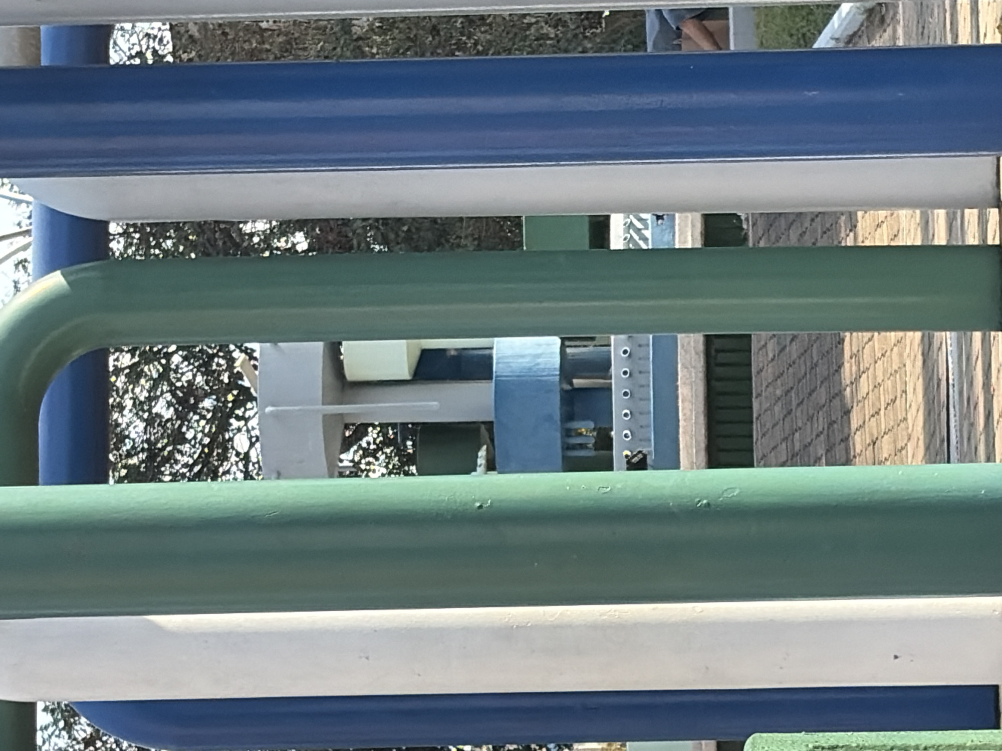 The image, taken in Guayana City, Venezuela, features a close-up view of a colorful outdoor structure, likely a playground, presented with a 90-degree clockwise rotation from its original capture. When viewed with correct orientation, prominent parallel tubular bars run horizontally across the frame, alternating in color between blue, white, and green.
Through the spaces between these bars, other parts of the playground and its surroundings are visible. To the left and behind the bars, there are glimpses of green foliage from trees, indicating an outdoor setting. On the right side, elements of the playground equipment can be discerned, including a light blue or gray section that appears to be part of a slide or ramp, along with white support posts. A distinctive vertical element with a series of small, uniformly spaced circular holes, possibly a ladder or a support beam with attachment points, is also visible. Further back, a textured wall or fence with a diagonal pattern, possibly made of wood, can be seen.
In the upper right portion of the original image (which would be the top right if viewed upright), a person's legs and feet are partially visible, suggesting someone is actively using or resting on the playground equipment. The scene is illuminated by bright, natural sunlight, indicating it is daytime with clear weather conditions. There is no visible text in the image.