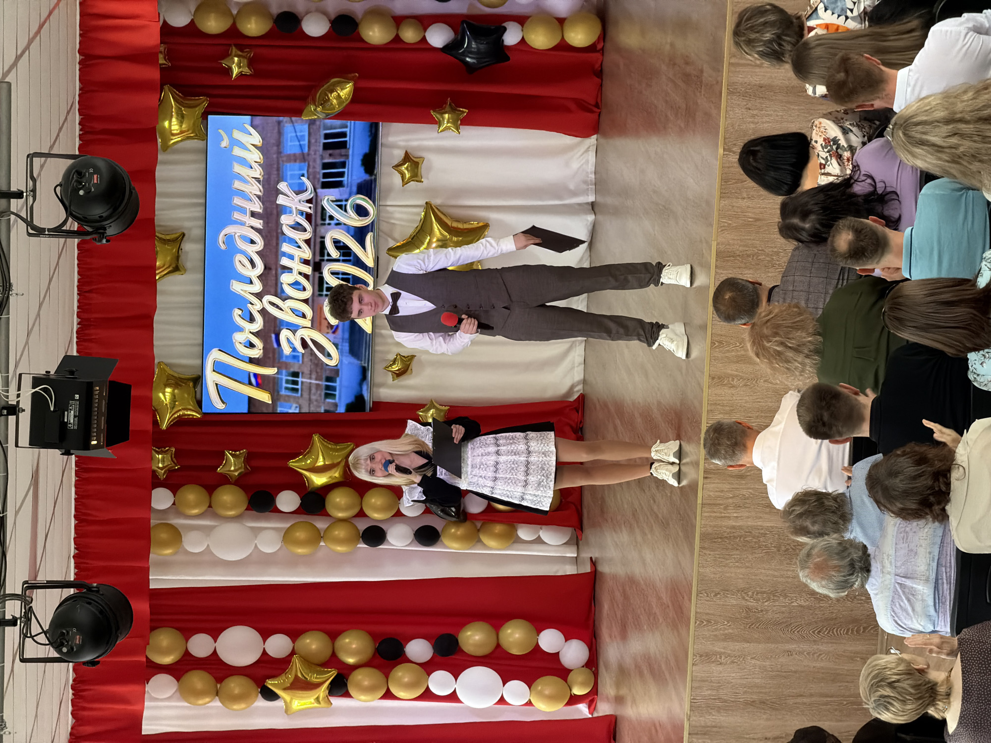 **Scene Description:**
The image depicts a staged event, likely a graduation or school-related ceremony, in what appears to be a hall or auditorium.
**People:**
* Two young people are standing on stage. The male student is wearing a gray suit, white shirt, a bowtie, sneakers, and holding a microphone and a black document. The female student is dressed in a black and white dress, has blonde hair, and is holding a microphone and a black folder.
* There's an audience comprised of people, with focus on their heads and shoulders, in the background, presumably watching the presentation.
**Subjects and Objects:**
* **Stage Decorations:** The stage is decorated with alternating vertical red and white drapes. A screen displays the Cyrillic text "Последний звонок 2026" (Last Bell 2026). Gold, white, and black balloons are arranged along the top of the drapes. Gold star-shaped balloons also adorn the stage.
* **Lighting:** There are theatrical stage lights on stands to the left side of the stage.
* **Flooring:** The floor appears to be wooden.
**Actions:**
* The two young people are presenting or performing on stage.
**Location:**
* The city of Izhevsk, Russia, is not directly determinable from the image.