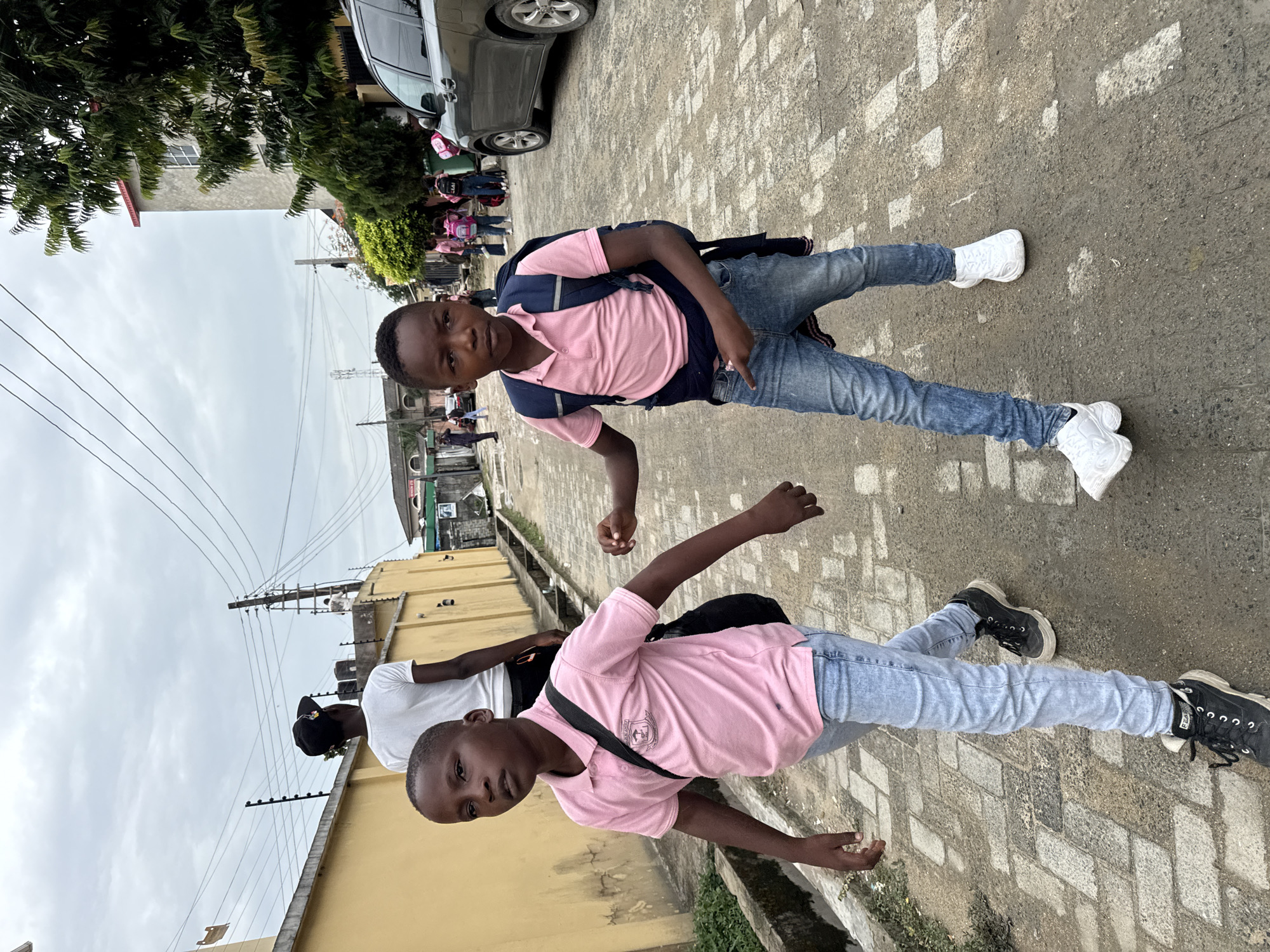 Two boys are standing on a brick-paved street with a building on the left and cars on the right. The boy in the foreground wears a pink shirt, light blue jeans, and black shoes and a black backpack with straps around his shoulders. The boy in the background is wearing a similar pink shirt with navy blue accents, jeans, and white shoes, and also has a backpack on. There is a man partially visible behind the boy in the foreground, wearing a white shirt and a black hat. Other people are standing near the vehicles to the right in the background. Ado, Nigeria.