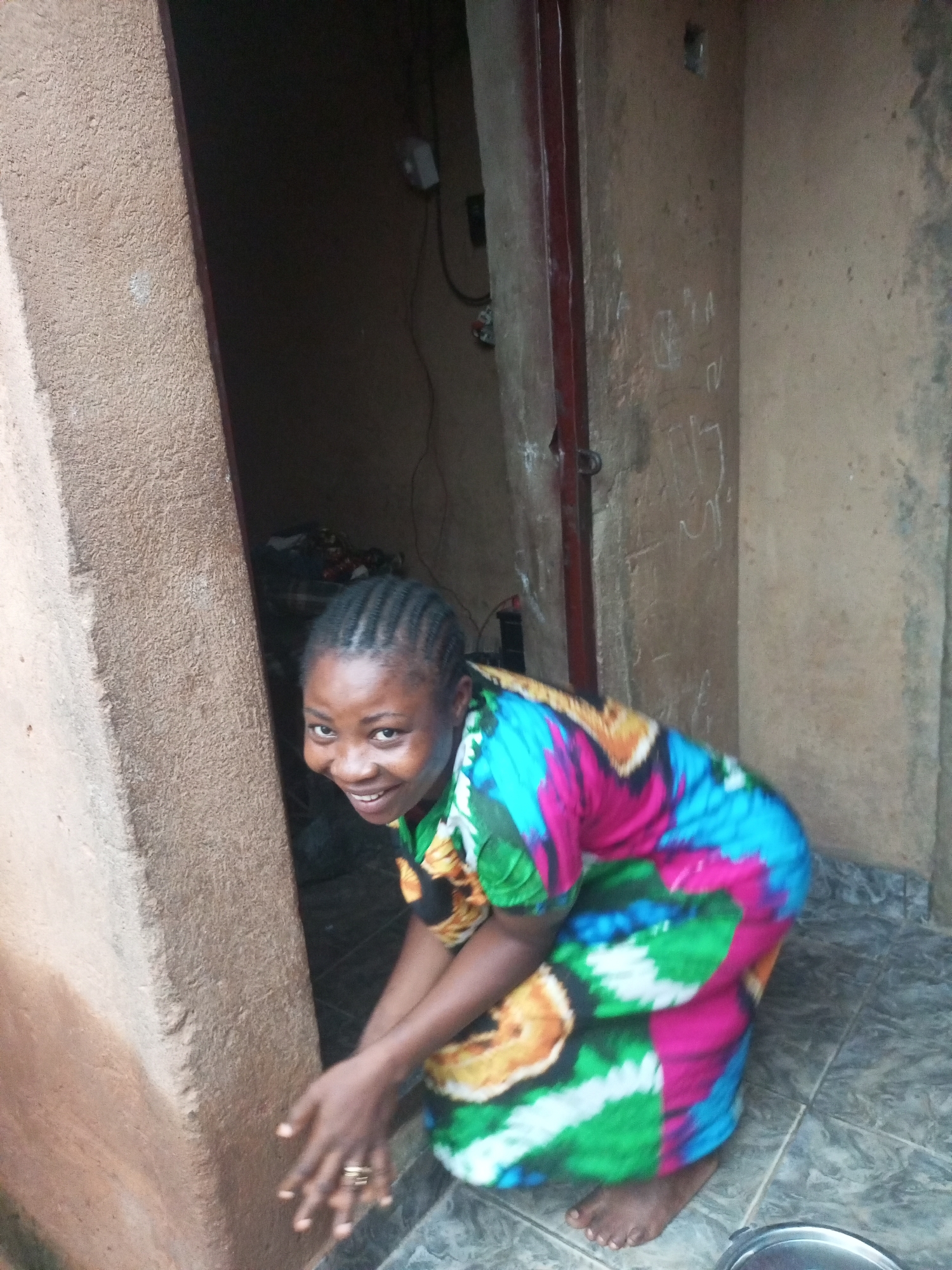 A young Black woman with braided hair and a colorful dress kneels in a doorway. Her head is turned to the left, and she is smiling at the camera. Her hands are clasped in front of her. The background is a textured, tan wall and a dark doorway. The floor is tiled. The image appears to be taken outdoors in a rustic setting, likely in a residential area within Tempure, Nigeria.