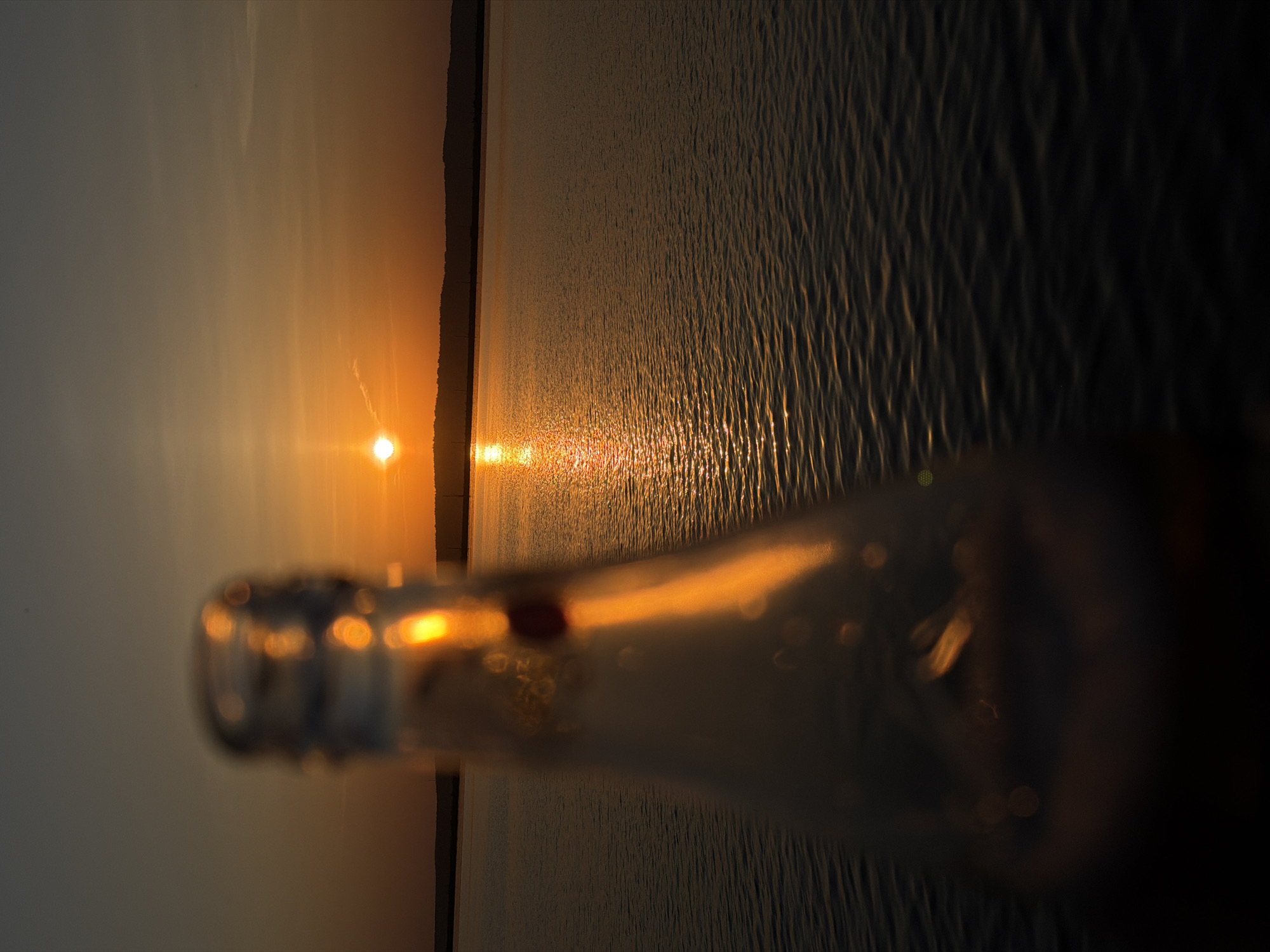 A blurry image oriented sideways, showing a sunset over water, with a bottle partially obstructing the view. The sun is a bright orange orb on the horizon, with a line of reflected light stretching across the water towards the viewer. Silhouetted land is visible on the horizon beneath the sun. A clear glass bottle is prominently featured in the foreground, partially out of focus. The light from the sunset is refracted and distorted through the glass, creating a series of bright highlights and blurry reflections within the bottle's contours. Based on the presence of the Votkinsk Reservoir, this is likely the city of Chaikovsky, Russia.