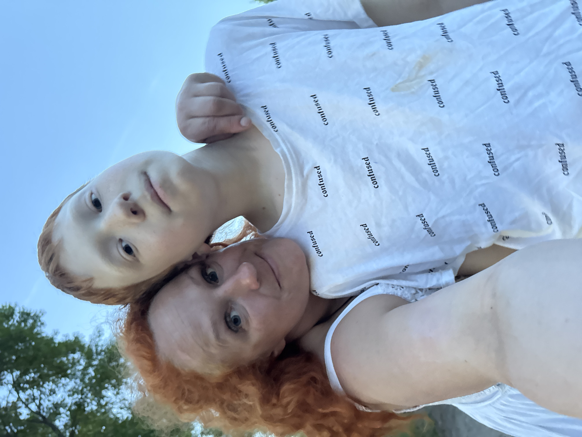 Here is a forensic description of the image:
**Scene:** A low-angle selfie featuring a woman and a child, likely taken outdoors based on the sky and trees visible in the background. The image is oriented with the subjects tilted to the side.
**Subjects:**
* **Woman:** An adult female with red, curly hair. She is wearing a white tank top and is positioned closer to the camera, smiling.
* **Child:** Appears to be a fair-skinned child with red hair. The child is wearing a white t-shirt printed with the word "confused" repeated. The child's head is positioned above and to the side of the woman's.
**Objects:**
* White t-shirt with the word "confused" printed repeatedly.
* White tank top worn by the woman.
* Sky visible in the upper portion of the image.
* Trees partially visible in the lower left corner.
**Actions/Interactions:** The subjects are posing for a selfie. The child is touching the woman's shirt near the neckline.
**Environment:** The setting appears to be outdoors, with the presence of trees suggesting a park or green space.
**Location:** The city of Chaikovsky, Russia is not discernable from the image.
