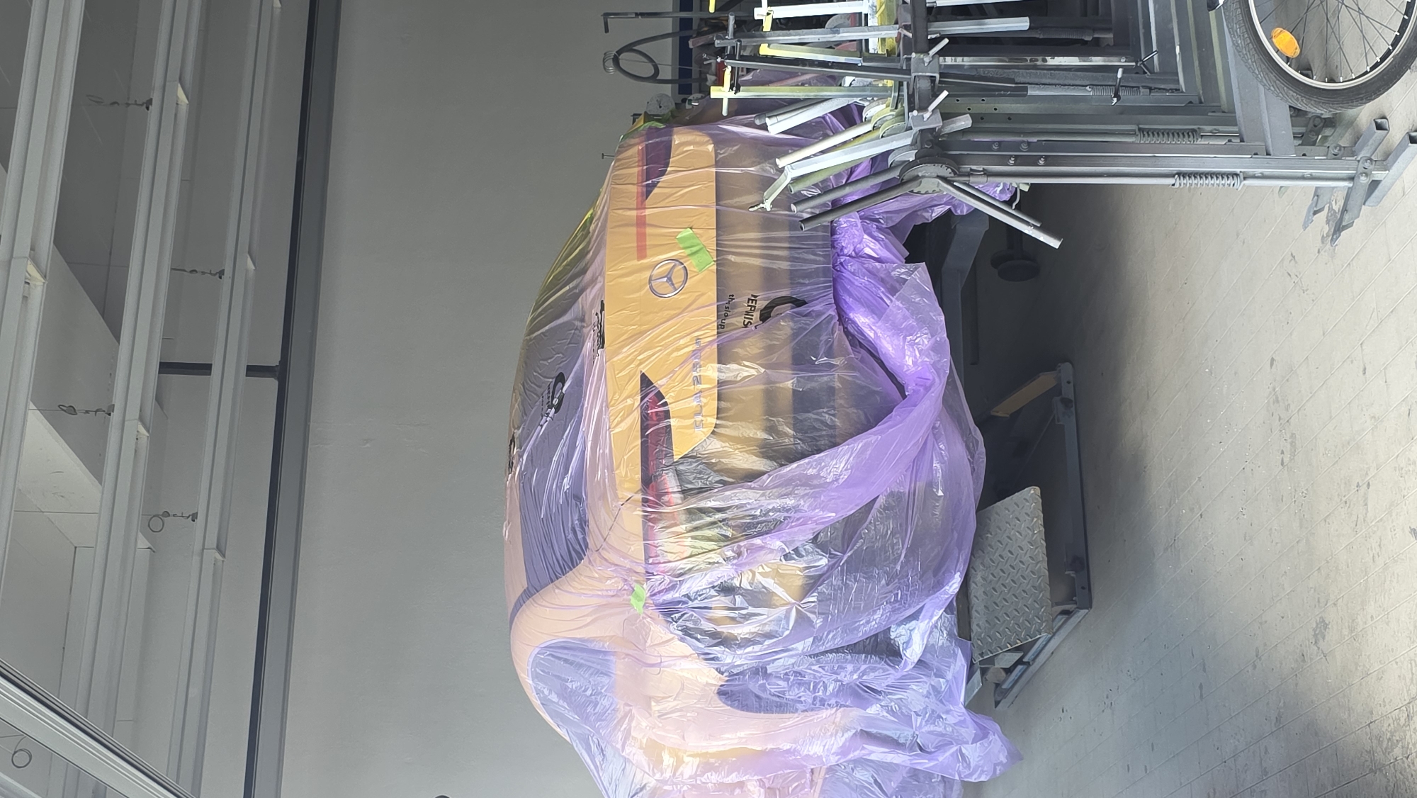 An orange car is covered in purple plastic wrap in a garage. The car is on a lift and its rear is visible. The Mercedes-Benz logo is visible on the back of the car. There are some metal racks and a bicycle in the foreground.