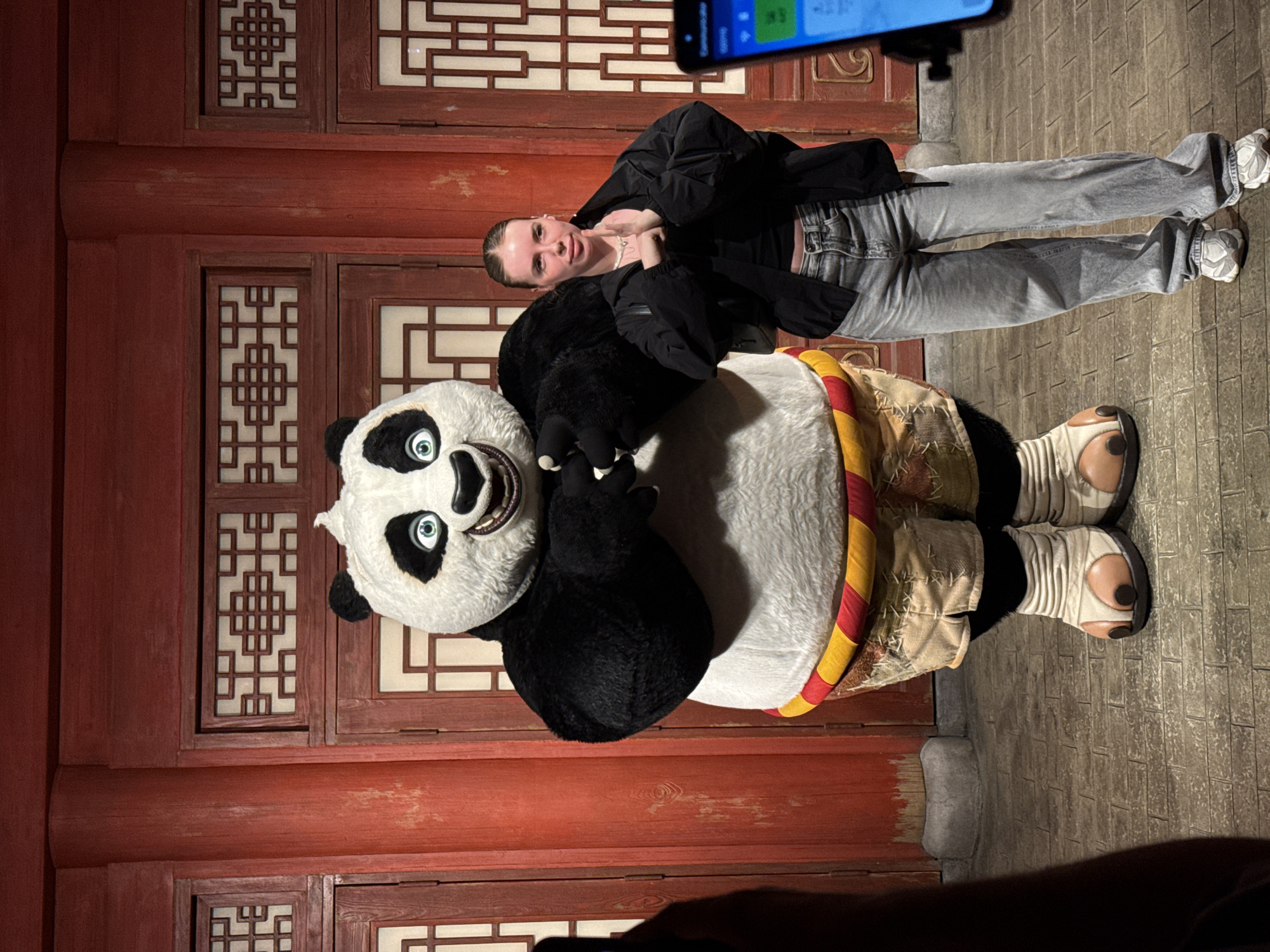 A woman poses with a Kung Fu Panda character costume in what appears to be a staged or themed setting. The woman is light-skinned with her hair pulled back, wearing a black jacket over a black top, grey jeans, and light-colored sneakers. She has one hand raised to her chest and the other touching the costume. The Kung Fu Panda character is standing next to her, fully costumed with a white and black panda head, black plush paws, and a costume resembling traditional clothing with a yellow and red rope belt.
The background consists of a traditional-looking Chinese-style wooden structure with intricate lattice window designs and red painted walls. The floor is paved with brick. The scene is illuminated, suggesting it is indoors or in a well-lit area.
Given the architectural style and thematic elements, the location is highly likely to be in the Tongzhou District, China.