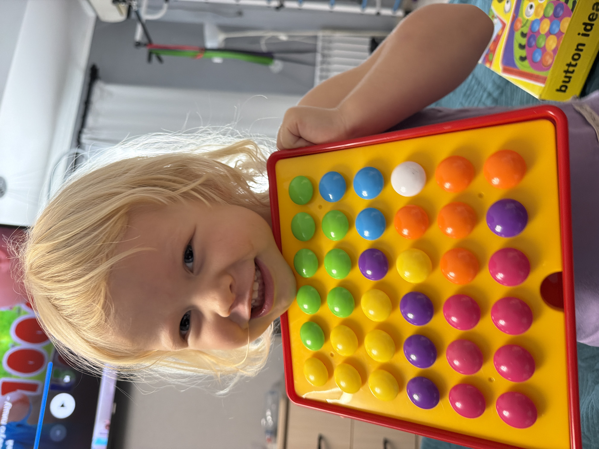 A close-up, medium-angle shot shows a young, fair-skinned, blonde-haired girl, around three to five years old, smiling slightly with her head tilted to the left. She is holding a yellow and red rectangular toy with a grid of colorful, round buttons in rows of blue, white, orange, purple, green, and pink. A book with the partial title "button idea" is visible to the right of the toy. The girl is wearing a purple t-shirt. In the background, a television screen, a laundry rack, curtains, and a dresser are present. The location is not identifiable as Sochi, Russia.