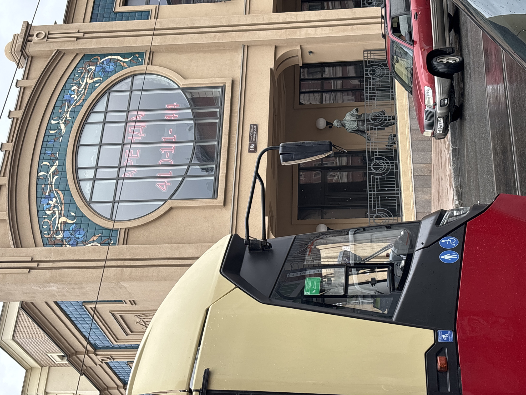 The image shows a street scene in Nizhny Novgorod, Russia, likely on Bolshaya Pokrovskaya Street, given the street sign visible on the building. In the foreground, a portion of a red and yellow tram is visible, its driver's side in focus. On the tram's lower exterior, there are stickers indicating "ATTENTION VIDEO SURVEILLANCE" and icons for pedestrians and wheelchair users.
Behind the tram, a building with a beige facade and ornate detailing stands. A large, arched window dominates the upper portion of the building, featuring a decorative tiled pattern with blue and brown elements. Inside the window, neon pink text reads "АРЕНДА" (rent) and a phone number "410-11-44". Below this large window, a street sign reads "18а улица БОЛЬШАЯ ПОКРОВСКАЯ" (18a Bolshaya Pokrovskaya Street).
On a balcony in front of the building, a statue of a person is visible, along with a railing. To the right of the building, a red Subaru Forester SUV is parked on the street. The street appears wet, suggesting it may have recently rained. The sky is overcast.