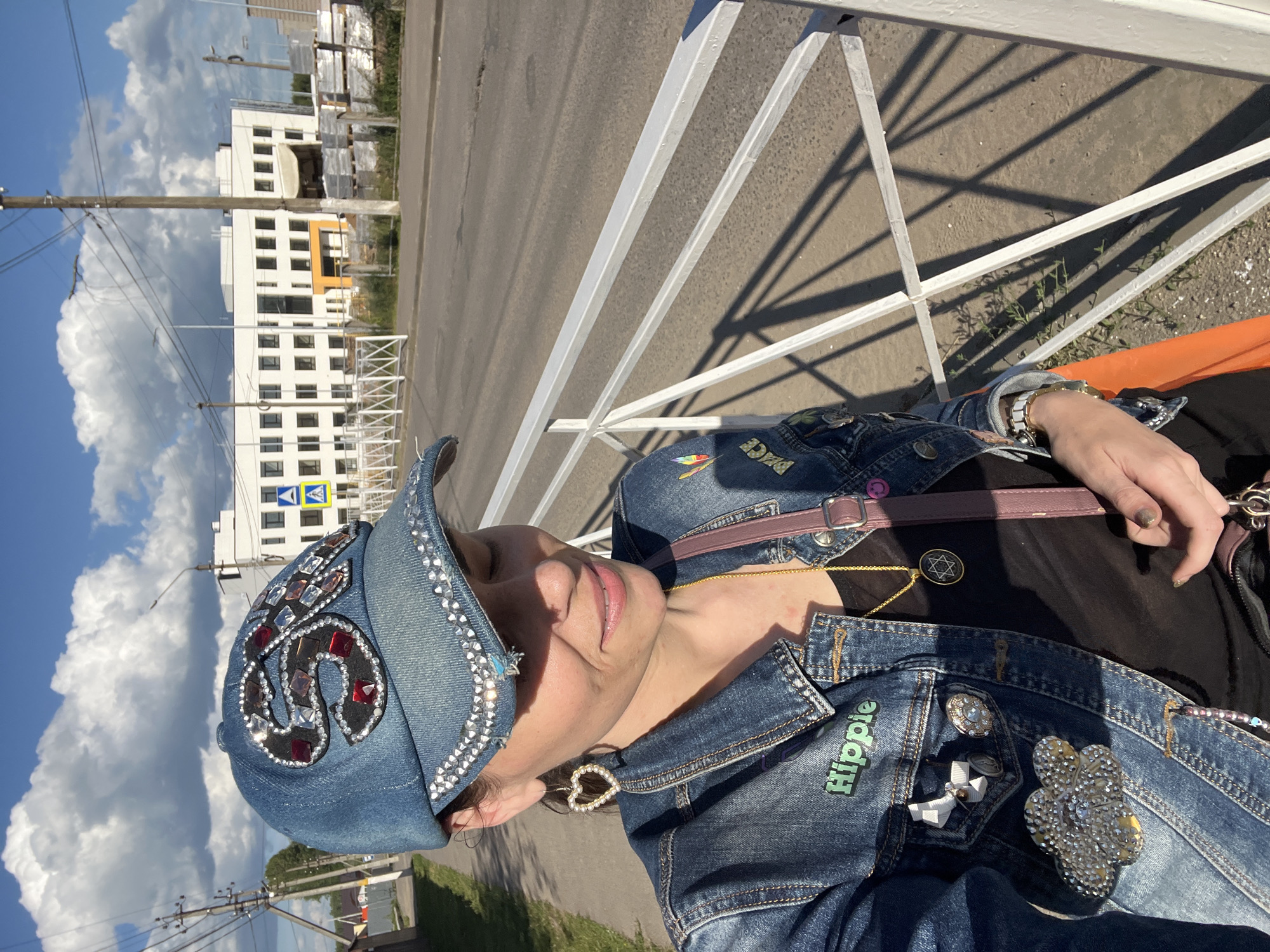 A woman is captured in a close-up, sideways shot, wearing a denim jacket and a decorated denim baseball cap. The jacket is adorned with several pins, including one that reads "Hippie" in green lettering, a floral brooch, and a small white bow. She wears a gold necklace with a Star of David pendant and a watch with a white band. The cap is also embellished with rhinestones and red and black accents. A portion of an orange surface and a purple strap from a bag are visible. The background shows a paved road, a white metal fence, and a large white building with many windows. A bright blue sky with fluffy white clouds suggests it is daytime with fair weather. The woman's eyes are closed, and her head is tilted back, giving the impression of relaxation or enjoyment in the sunlight. The scene appears to be outdoors, possibly near a street or walkway given the road and building. The overall mood is casual and sunny.
