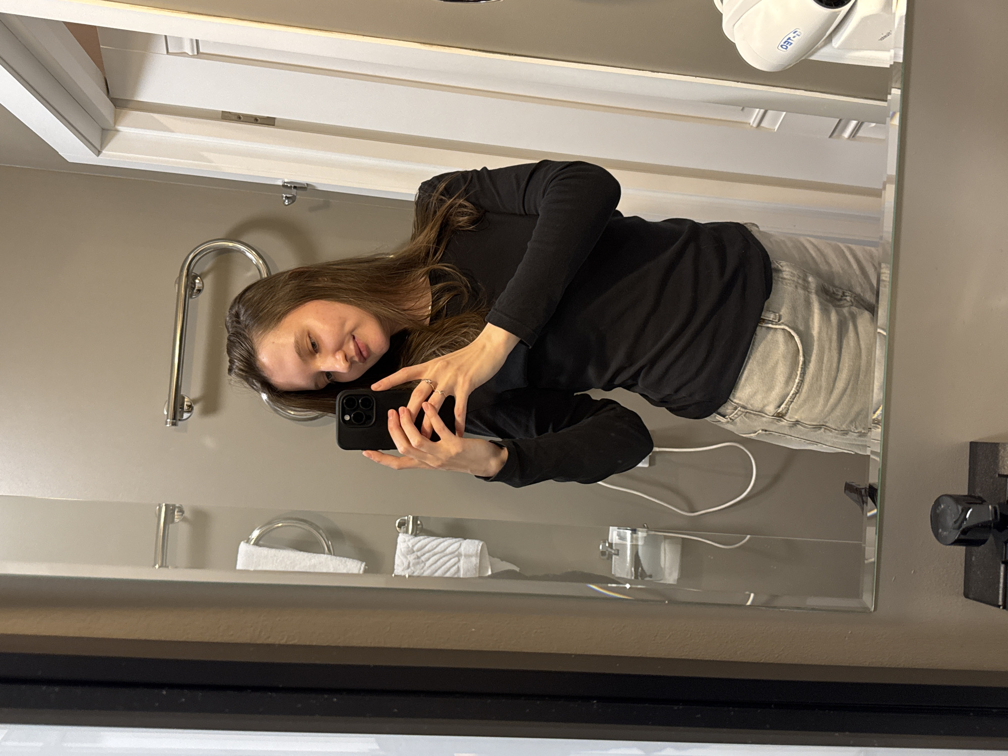 A woman is taking a mirror selfie in a hotel bathroom. She is wearing a black long-sleeved shirt and light-colored denim shorts. Her hair is long and brown. She is holding a black smartphone in her right hand, and her left hand is resting on her hip. The bathroom has gray walls and a large mirror. There are towels on a rack and a toilet paper dispenser visible in the mirror's reflection. A hairdryer is mounted on the wall to the right of the mirror. The overall scene suggests a private, domestic setting.