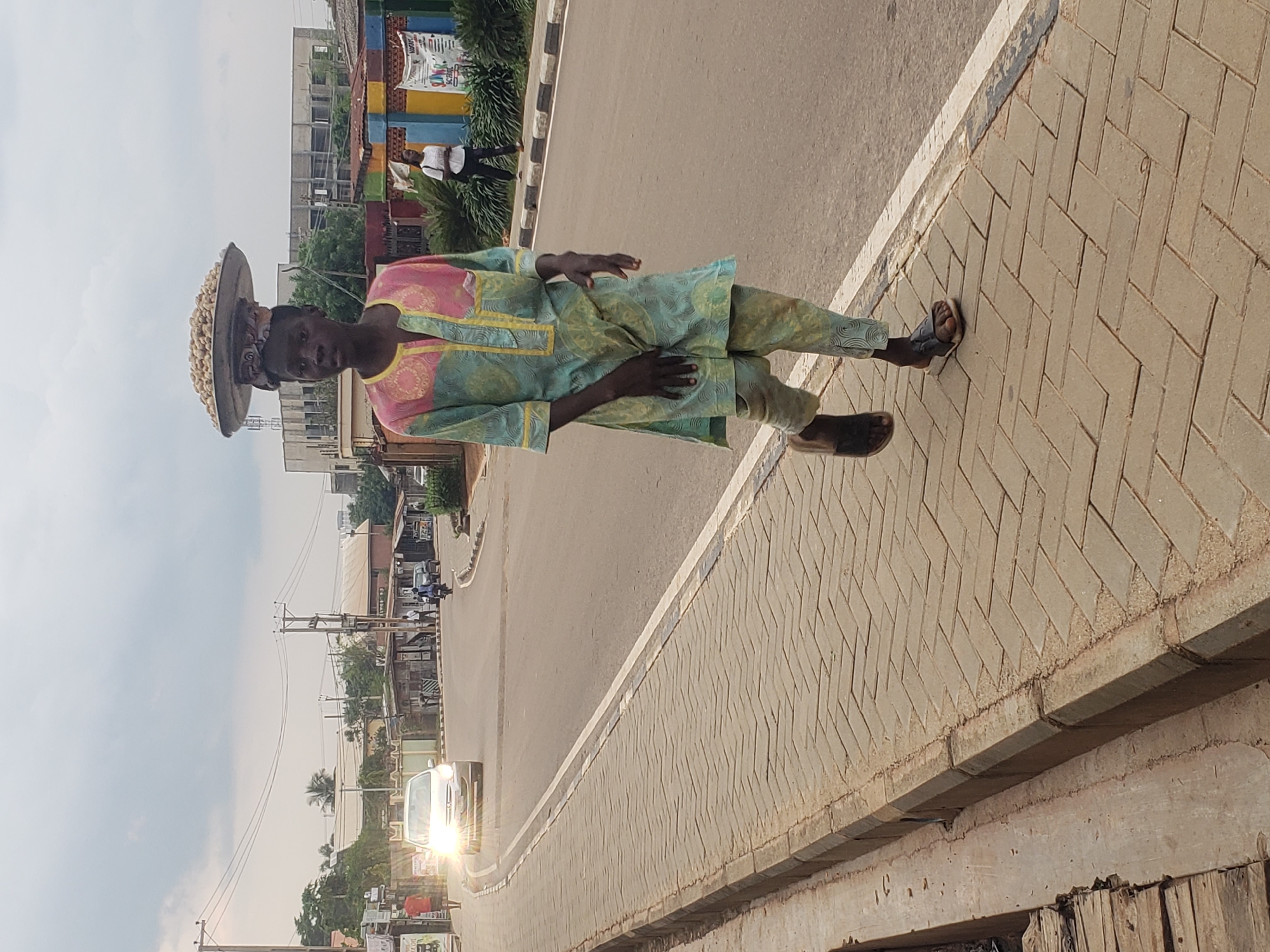 A young African person wearing a patterned dashiki and trousers walks along a paved sidewalk next to a road in Narayi, Nigeria. The person is balancing a large metal tray filled with nuts on their head. The sidewalk is made of interlocking bricks with a concrete curb. The road is asphalt with a white line marking the edge. In the background, there are buildings, power lines, and other pedestrians. A silver car with its headlights on is approaching on the road. The sky is overcast.