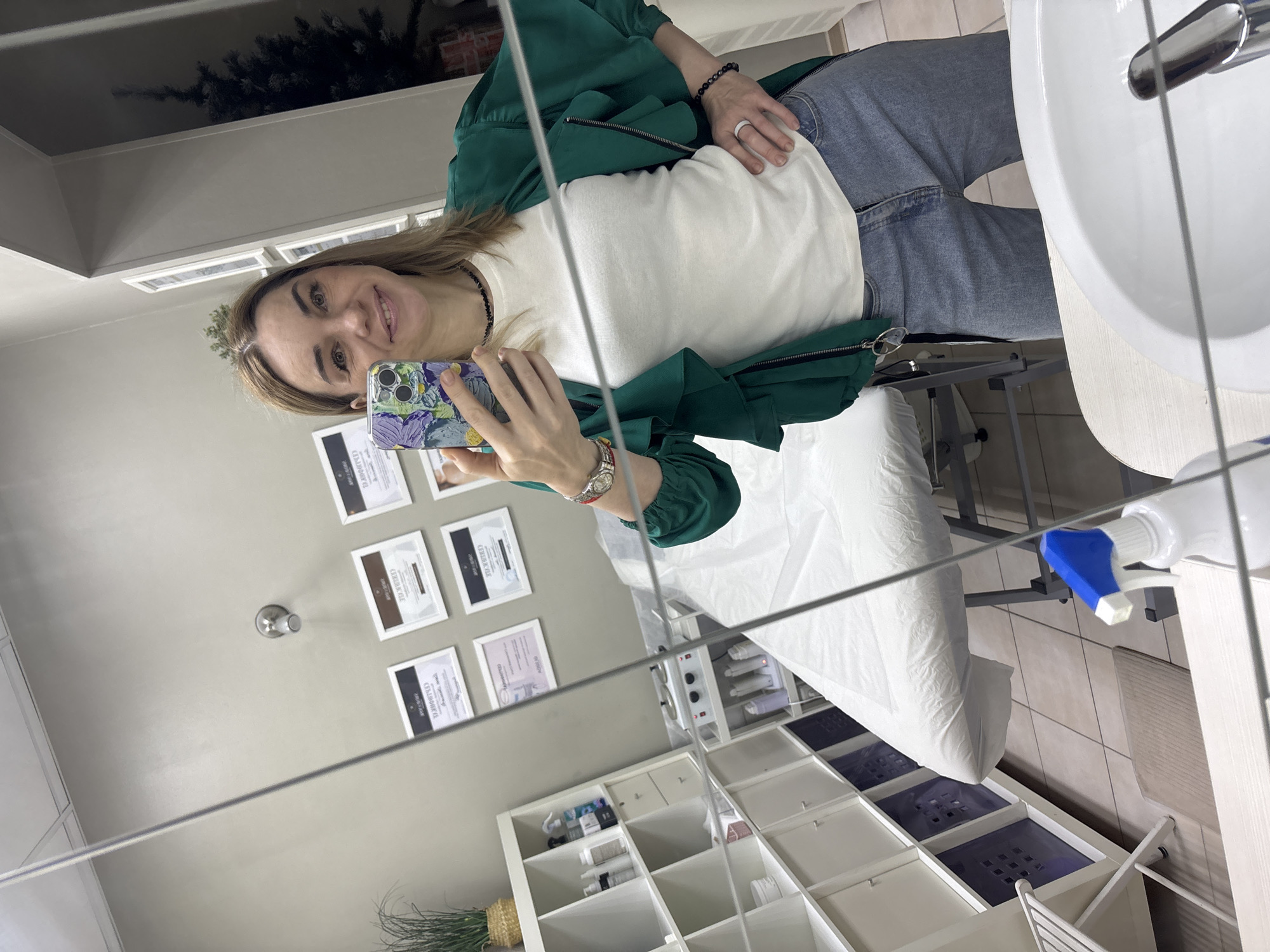 A woman is captured in a mirror selfie, smiling broadly at the camera. She has medium-length brown hair and is wearing a white t-shirt, a dark green jacket, and blue jeans. She holds a smartphone with a colorful, possibly floral, case. Her left hand displays a black beaded bracelet and a white ring, while her right wrist has a silver watch with a red band.
The scene reflects a professional indoor setting, likely a beauty salon, spa, or clinic. The walls are light grey, and the floor is tiled. To the right, a white sink with a chrome faucet is visible. Below the woman and extending to the left, there is a white treatment bed or couch covered with a clean white sheet. Below the bed, a white shelving unit holds numerous bottles and containers, suggesting beauty or treatment products. A spray bottle with a blue nozzle is positioned near the sink.
On the wall behind the woman, several framed certificates are displayed. The text on these certificates is primarily in Cyrillic, with "СЕРТИФИКАТ" (Certificate) and "ОБ ОКОНЧАНИИ КУРСА" (On Completion of the Course) being visible on some, indicating professional qualifications. A small potted plant in a woven basket rests on the floor in the lower left, and a wall-mounted light fixture is above the certificates. The time of day is not apparent, and the environment appears well-lit by artificial light. The woman's expression conveys happiness and comfort.