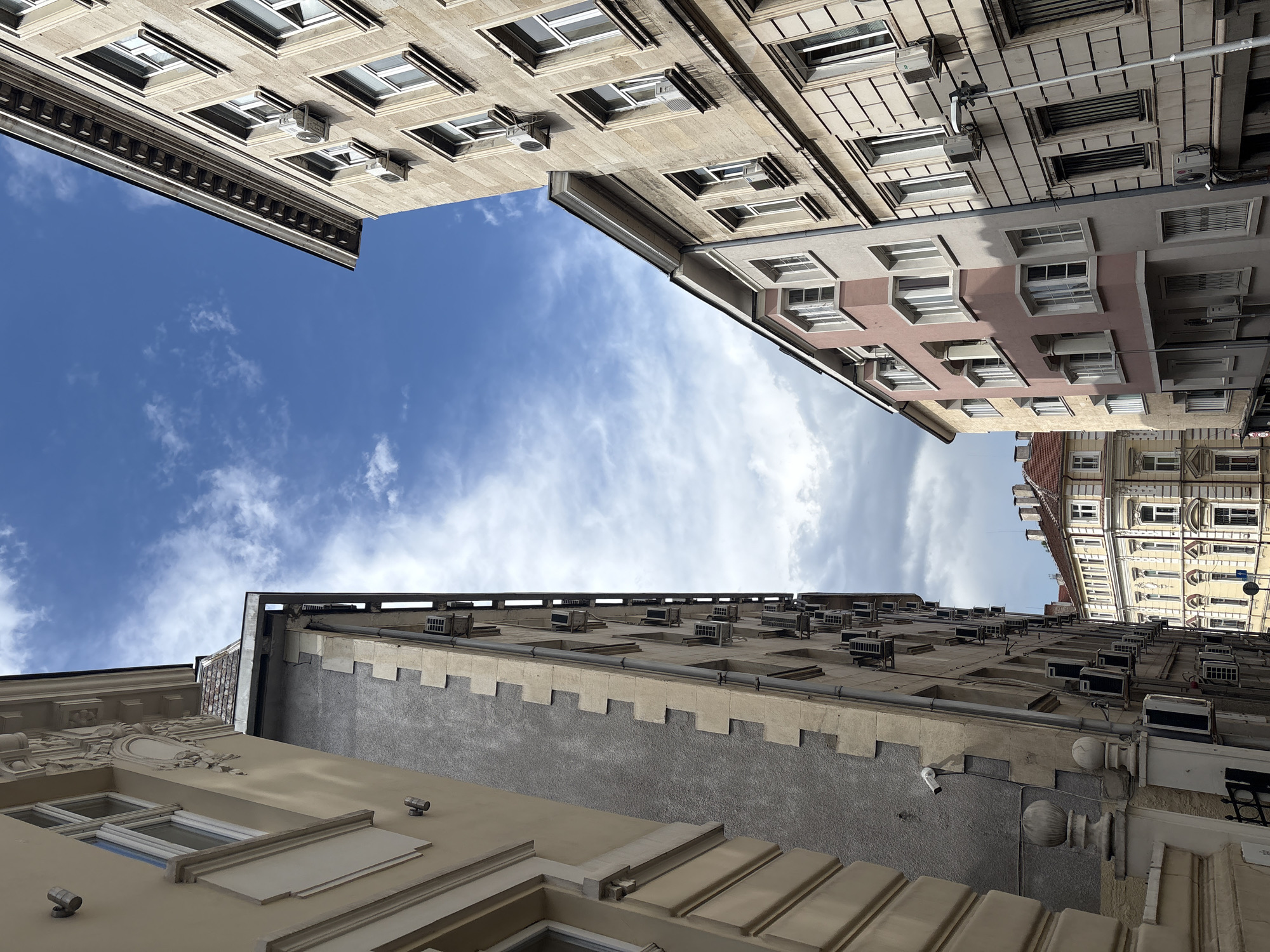 The image captures a dramatic worm's-eye view, looking upwards between several multi-story buildings towards a bright blue sky with scattered white clouds. The scene is presented with a 90-degree clockwise rotation from a typical upright perspective, giving the sky a vertical strip appearance in the center.
Dominating the central vertical expanse is a clear blue sky, adorned with soft, wispy white clouds. The bright illumination suggests it is daytime, likely mid-day, with the sun high above.
Buildings flank all sides of the sky. To the top-left (left side of the image as viewed), a light-colored building, possibly beige or light grey, displays multiple rectangular windows and numerous compact air conditioning units mounted on its facade. A decorative cornice and horizontal banding provide architectural detail.
To the top-right (right side of the image as viewed), another multi-story building features a varied facade, with sections in a light beige and a muted pink/terracotta color. This building also has many windows and air conditioners. In the distant background, a building with a red-tiled roof and an ornate, yellowish facade can be seen.
The building extending across the bottom (left side in the provided original image) appears closest and is quite prominent. It features a textured, dark grey lower section, contrasting with a lighter, smoother upper section that boasts a decorative, crenellated-like pattern along its top edge. Many air conditioning units are visible on its upper facade, and ornate architectural elements, including decorative moldings, a security camera, and two small spotlights, are present on its lower visible part.
The overall architectural style suggests an urban European setting, consistent with Sofia, Bulgaria, combining classical elements with the visible modern additions of air conditioning units on nearly every facade. There are no people visible, nor is any legible text apparent.