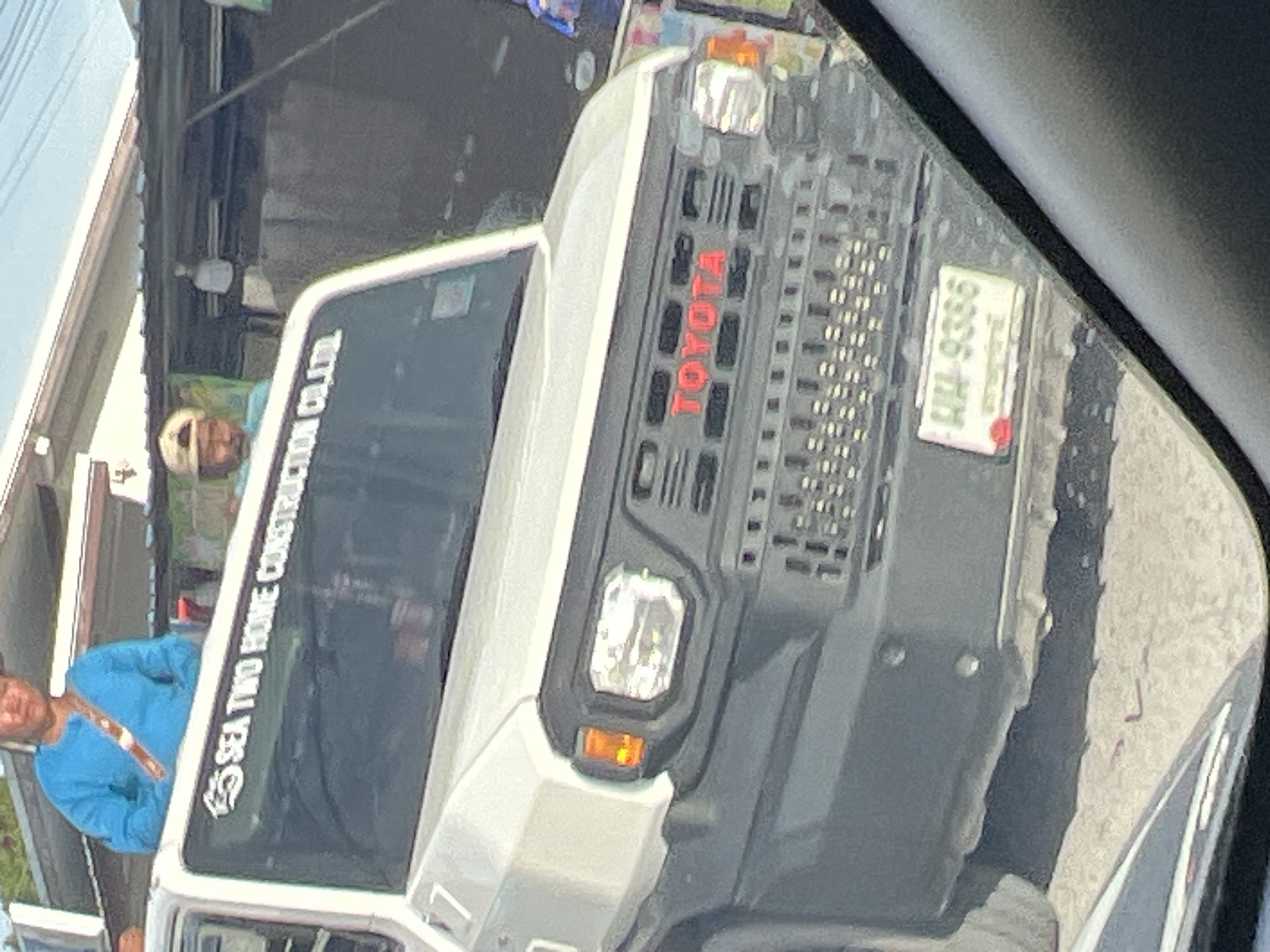 Captured through a side-view mirror, the image shows a portion of a white Toyota truck. The red "TOYOTA" logo is visible on the truck's front grille, along with a Thai license plate reading "HH-9386". The reflection also captures two individuals. One is wearing a blue shirt and a light-colored strap or lanyard, while the other is partially obscured, wearing a light-colored hat. Decal writing on the front windshield reads "SEA TWO HOME CONSTRUCTION CO.,LTD". The location, as stated, is Baan Bang Rak, Thailand.