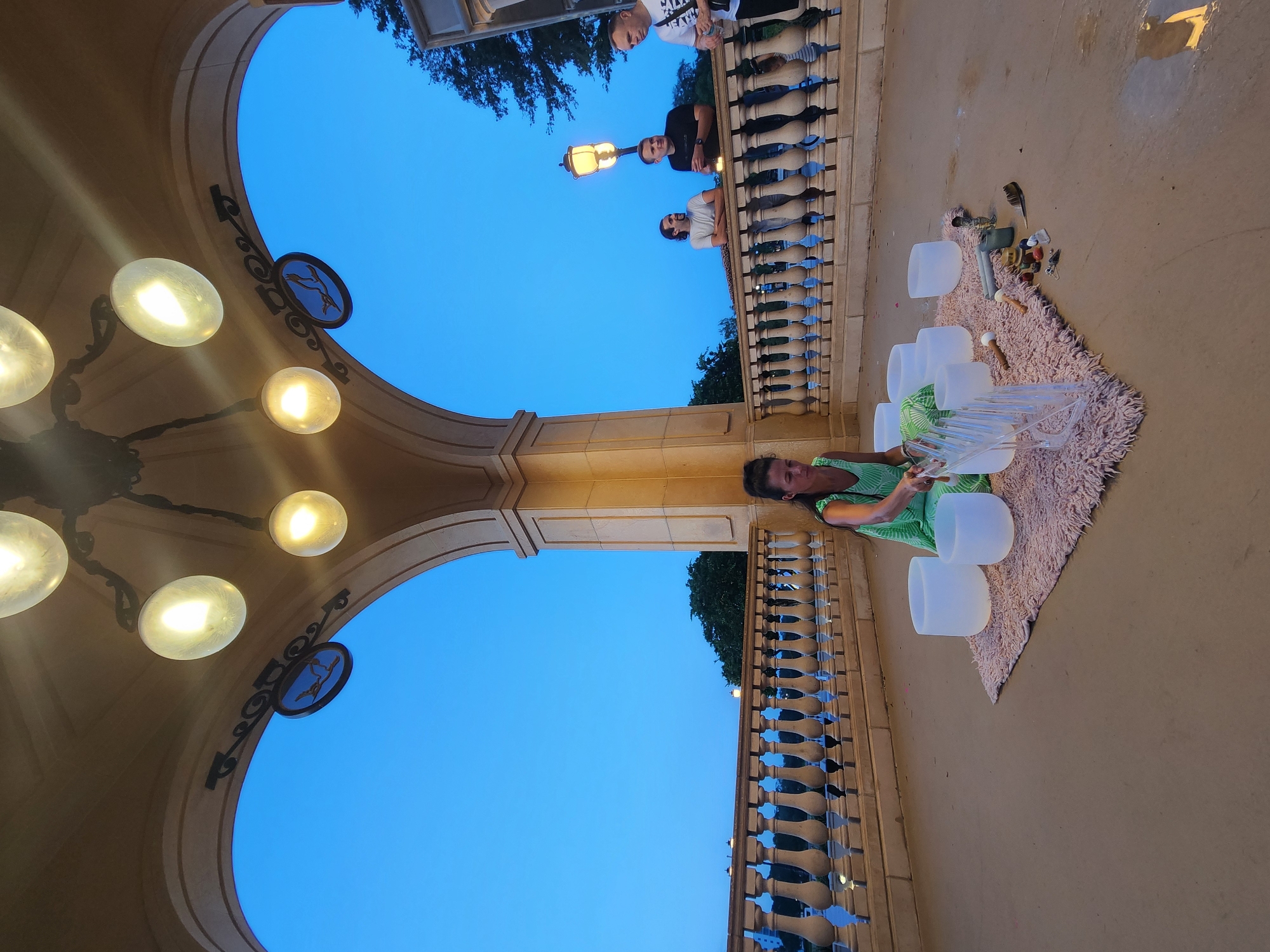 At dusk, a woman in a green dress sits on a pink rug in an open-air rotunda, surrounded by several large crystal singing bowls, some of which she is striking with mallets. Several people stand near a railing in the background, observing. The rotunda is built of tan stone and features ornate lamps hanging from the ceiling. The sky is a deep blue, indicating evening. This scene is taking place at the Cranes Roost Park in Altamonte Springs, Florida, United States of America, a popular venue for outdoor events and gatherings.