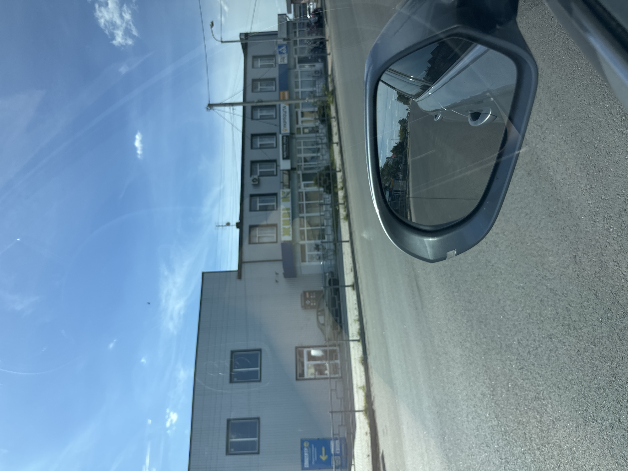 This is a vertical, eye-level shot of a street scene in Sarny, Ukraine, as seen from a vehicle. The main subjects are a modern building with several windows, a side-view mirror of a car, and the road. The building appears to be commercial, with signs visible on its facade. One sign is in Cyrillic and reads "УКРПОШТА" (UKRPOST), indicating a post office. The sky is a bright blue with scattered white clouds, suggesting it is daytime with fair weather. The road is paved with asphalt and shows a slight shimmer, possibly from the sun. The side-view mirror reflects the road and some greenery, offering a glimpse of the car's surroundings. The overall impression is that of a sunny day in a Ukrainian town, with the perspective suggesting the image was taken while driving. There are no people or animals clearly visible in the frame.
