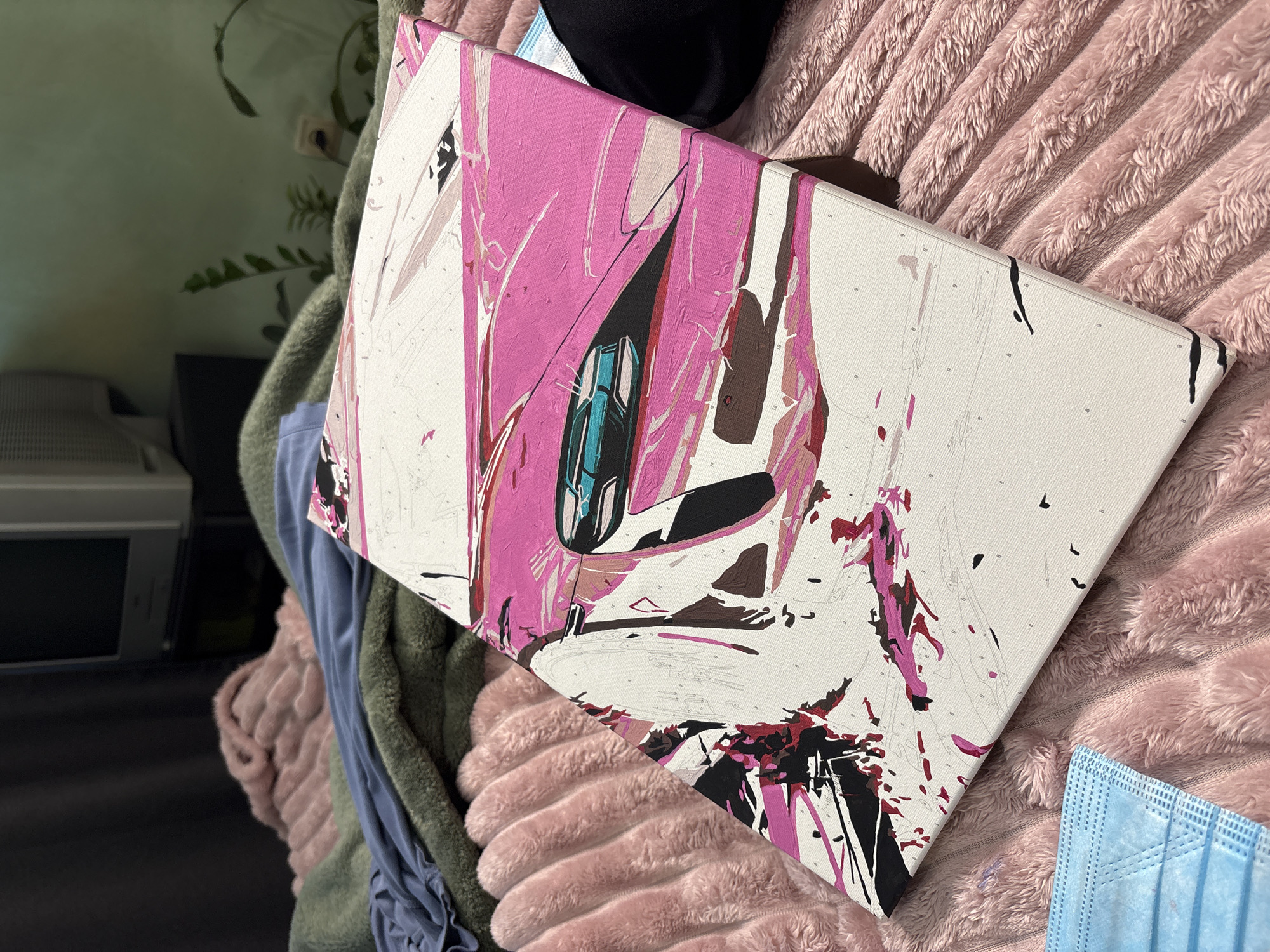 The image shows a partially completed paint-by-numbers canvas artwork resting on a pink, plush blanket. The artwork features a predominantly pink abstract design with white, brown, black, and teal accents. The canvas is positioned at an angle, revealing its white edges marked with numbered sections. In the background, there are glimpses of a light blue garment, a dark green throw, a white appliance or piece of furniture, and a potted plant. A blue surgical mask is visible in the lower right corner. The setting appears to be indoors. The location in the city of Unknown, Russia is not determinable from the image.