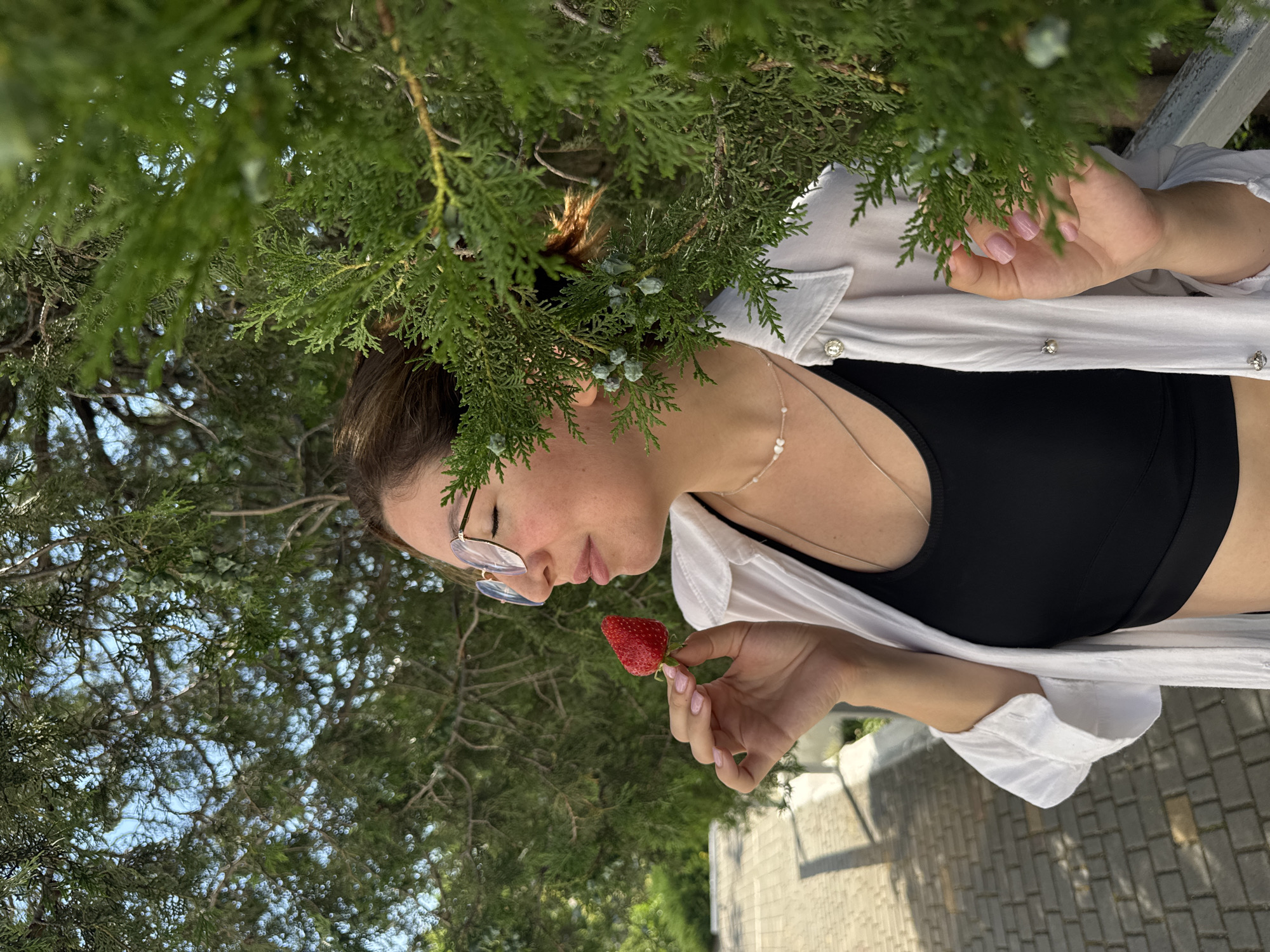 A woman with dark hair, wearing glasses, a white unbuttoned shirt over a black top, and necklaces, stands outdoors, holding a strawberry near her face with her eyes closed. She is surrounded by green foliage. In the background, a paved walkway is visible. The scene is likely in Odesa, Ukraine, given the context.