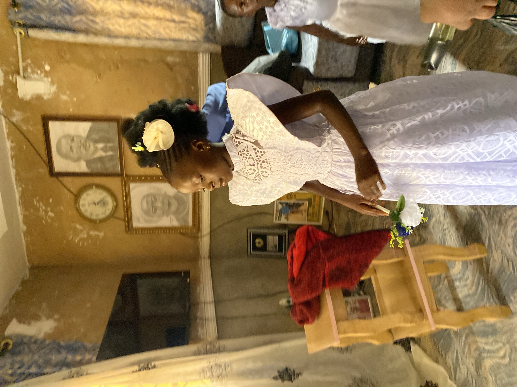 The image captures two girls in white dresses, likely communion dresses, indoors in a room that seems to be a residence in Kuru Dungus, Nigeria. The room is sparsely decorated with framed photos on the walls, including portraits, and a clock. One girl is prominently featured, and she is holding a small bouquet of flowers. A small wooden chair draped with a red cloth is also visible. The scene suggests a religious or celebratory occasion taking place within a modest home environment.