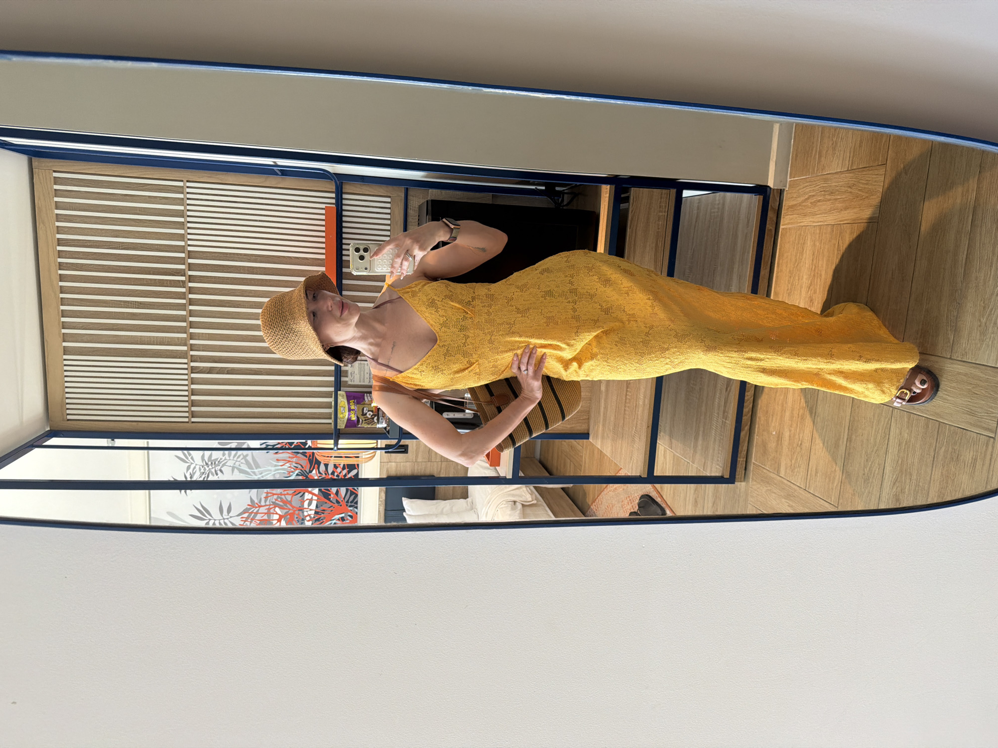 A woman stands in front of a large mirror, taking a selfie. She is wearing a yellow, floor-length lace dress, a straw hat, and brown sandals. A striped straw bag hangs from her shoulder. The mirror reflects a hotel room with a bed, a wooden shelving unit, and a wall decorated with vertical wooden slats. The flooring is light-colored wood. There is a framed artwork of coral and plants on the wall behind her. The scene is brightly lit, suggesting daytime. There is no indication of the location being in Dong Ninh Hoa Ward, Vietnam.