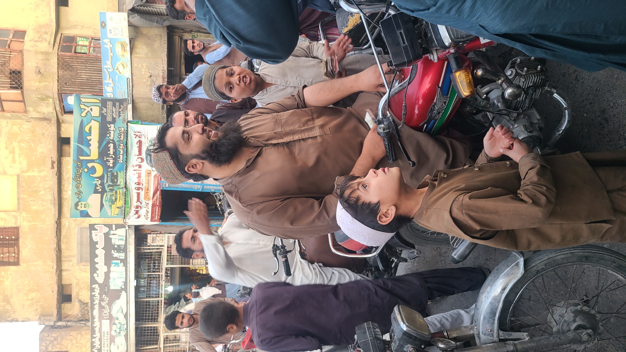 The image shows a crowded street scene in what appears to be Quetta City Tehsil, Pakistan. Multiple men and boys are gathered around several parked motorcycles. Some of the people are wearing traditional clothing, including caps. Shops with Urdu signage are visible in the background. The street is busy with activity, and the environment suggests a commercial or public space.