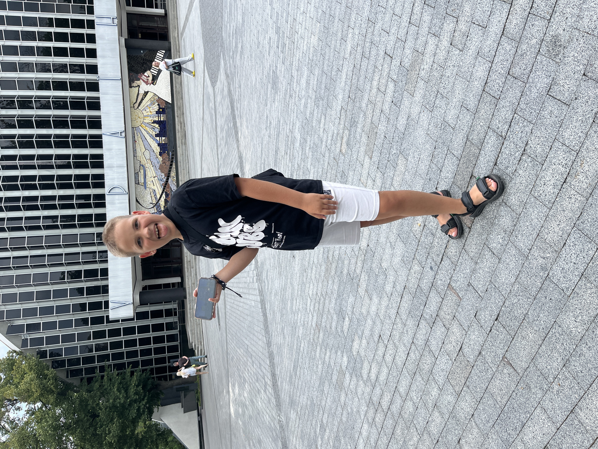 The image shows a young boy standing in an outdoor plaza in front of a large, modern building with a facade of black-tinted windows and a mosaic mural. The boy is wearing a black t-shirt with white lettering, white shorts, and black sandals. He is holding a smartphone in his left hand and sticking his tongue out at the camera. In the background, other people can be seen walking in the plaza and on steps leading up to the building. The ground is paved with grey cobblestones. The building appears to be the Kharkiv National Academic Opera and Ballet Theater, located in Kharkiv, Ukraine.