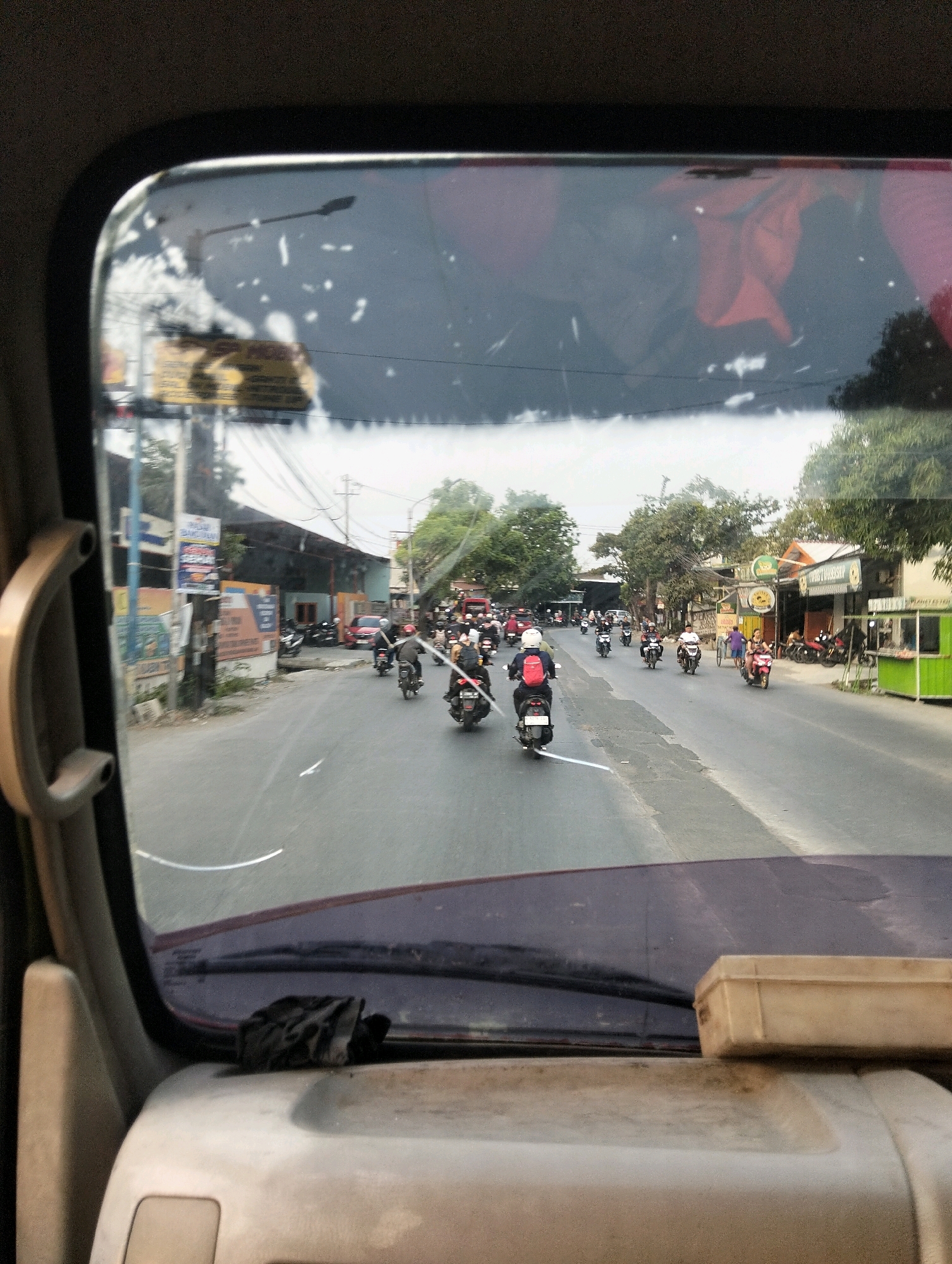 The image shows a street scene in Semarang, Indonesia, viewed from inside a vehicle, likely a car or van, through the windshield. The road is lined with buildings and trees. There is significant traffic, primarily consisting of motorcycles. Many of the motorcyclists are wearing helmets. Some buildings appear to be shops or small businesses, with signs visible on them. There are power lines and utility poles along the roadside. The sky is overcast. In the foreground inside the vehicle, a black cloth is resting on the dashboard. A tan-colored box is also visible on the dashboard. The windshield has some smudges or debris on it, affecting visibility in some areas. The overall impression is a busy street during daylight hours.