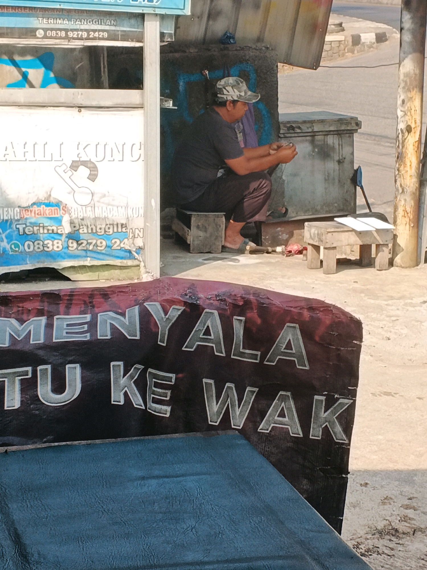 The image depicts an outdoor scene in Ungaran, Indonesia, likely on a street or sidewalk. In the foreground, a portion of a dark red banner with white text is visible, with the words "MENYALA" and "TU KE WAK" partially decipherable. A blue textured surface, possibly a tarp or seating, occupies the lower portion of the frame.
Further back, a man wearing a camouflage baseball cap, a dark t-shirt, and maroon pants is seated on a low wooden stool. He is hunched over, looking at something small in his hands. To his right, a light-colored concrete structure and a weathered wooden stool with two white sheets of paper on top are visible. Behind the man, a rough, textured wall with graffiti in blue and purple paint can be seen. The background suggests a street or paved area with some debris and a metal pole on the right side of the image. The lighting indicates it is daytime, with strong shadows suggesting sunlight. The overall impression is of a humble roadside establishment or waiting area.