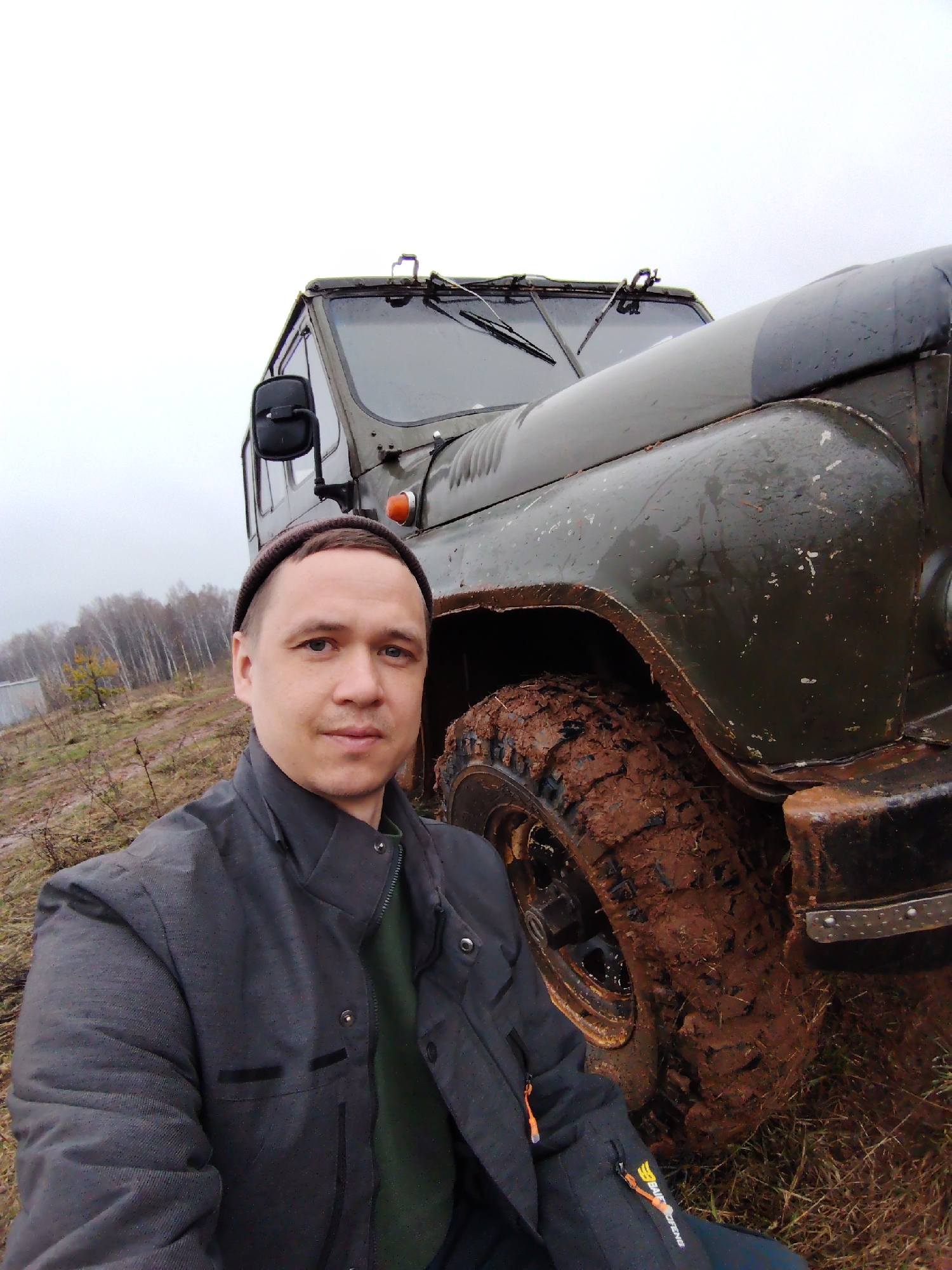 The image features a man taking a selfie outdoors in a rugged, muddy environment, likely in Марковский, Russia. In the foreground, a man in his 30s or 40s, wearing a dark grey jacket over a green shirt and a dark beanie, looks calmly into the camera.
To his right and slightly behind him is a large, dark green military-style truck, possibly a GAZ or UAZ model. The truck's front right fender and a heavily mud-caked, knobby tire are prominent, indicating recent off-road activity. The vehicle is splattered with reddish-brown mud, and its windshield and a side mirror are visible.
The background shows a sparse, rural landscape under an overcast sky. Bare deciduous trees line the horizon, suggesting late autumn or early spring. The ground is a mix of dry grass and mud. On the lower right sleeve or an attached item on the man's jacket, a visible logo reads "BAOFENG," a brand associated with two-way radios, accompanied by a stylized 'B'. The scene suggests an outdoor adventure or work in challenging, off-road conditions on a cloudy day.