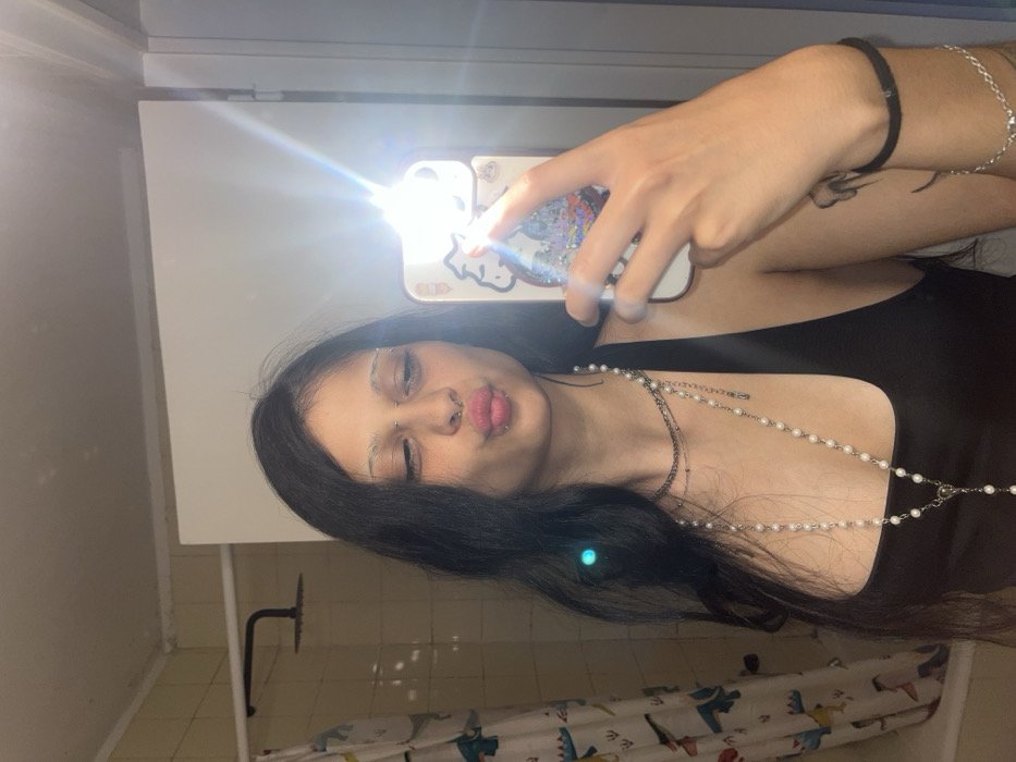 A woman in a black tank top takes a mirror selfie with a phone decorated with Hello Kitty. Her dark hair flows around her face, which features multiple piercings and light-colored eyebrows. She wears a beaded necklace and a shorter silver chain. On her wrist, she has a bracelet and a black elastic band. The bathroom background includes a shower curtain with cartoon characters and beige tiles. The phone's flash creates a glare in the photo.